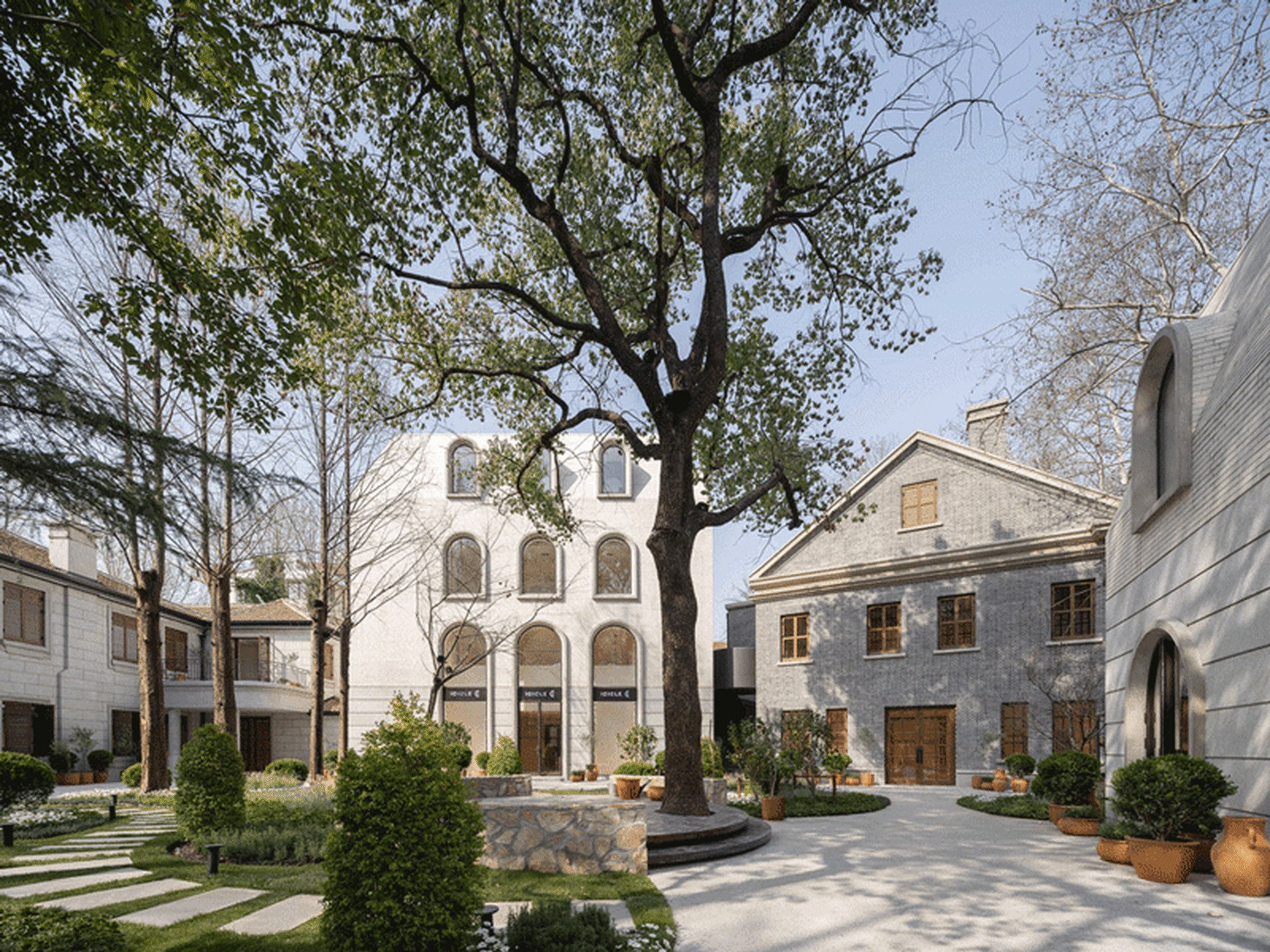 Modern building exterior featuring brown brick and white concrete materials, with a lush green courtyard and tall trees.