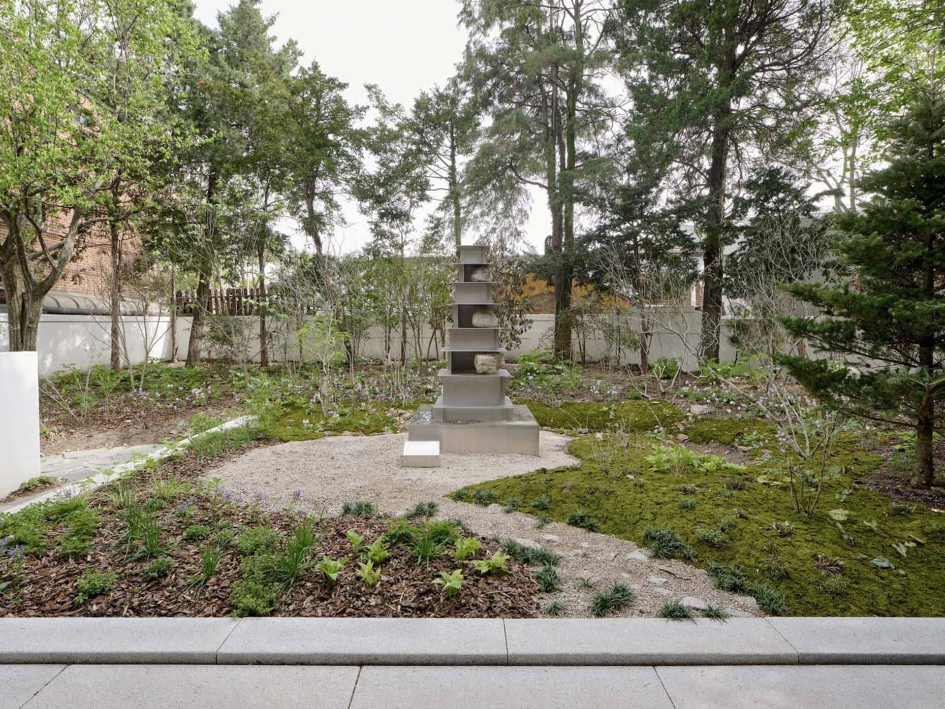 A contemporary garden with moss, trees, and a minimalist sculpture on a gravel path.
