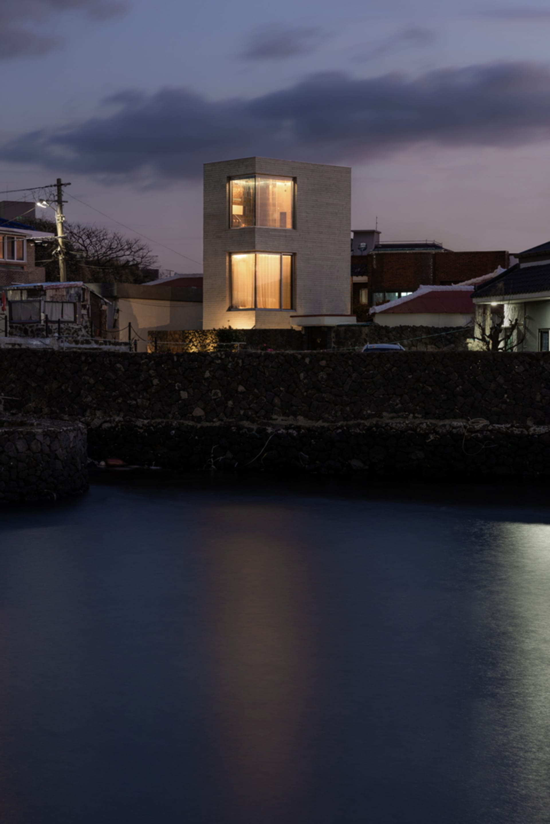 Modern waterfront house with concrete and glass exterior beside calm water during dusk