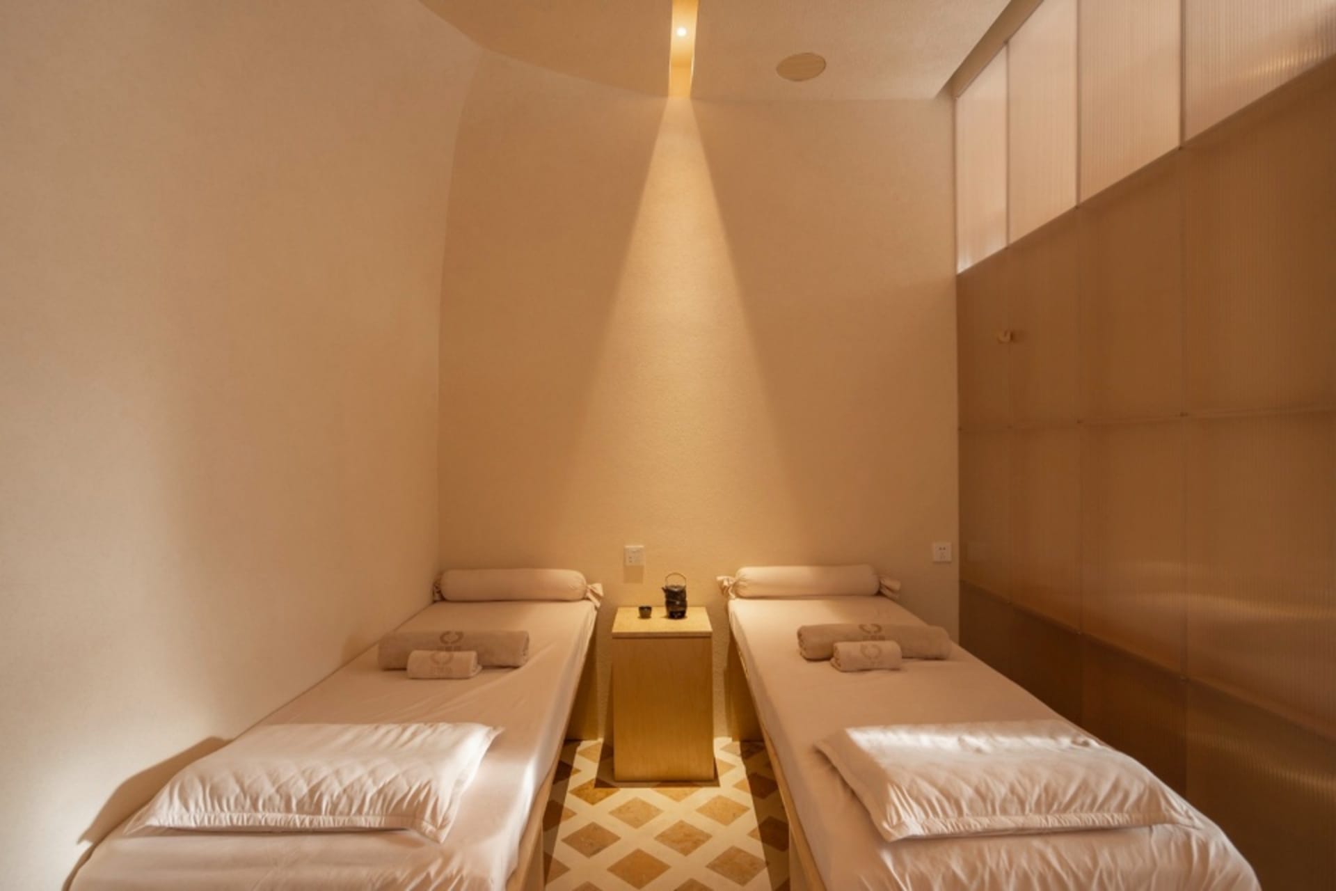 Two white spa beds with neutral-toned sheets and pillows, placed in a minimalist spa interior with warm lighting.
