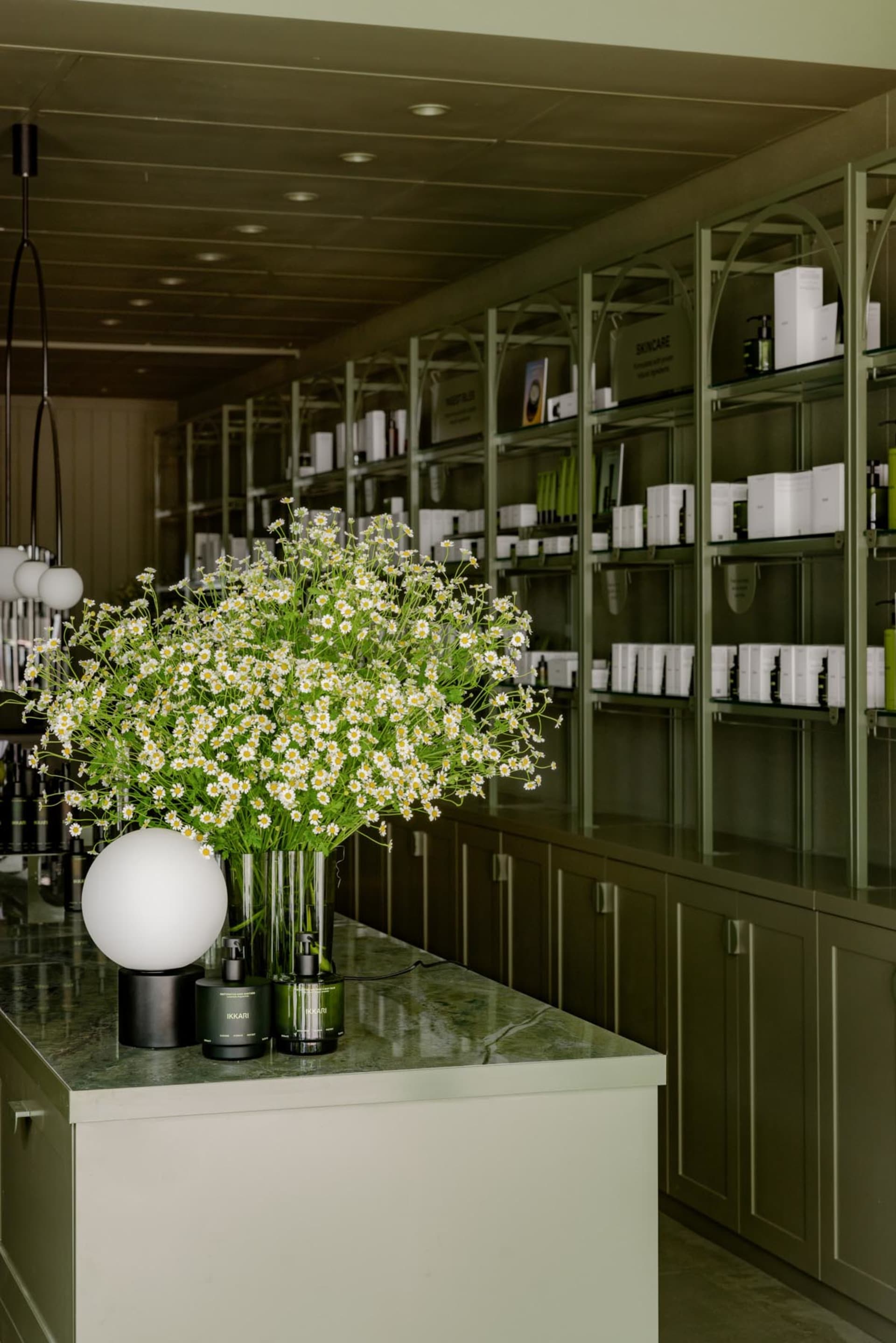 A spa interior with a large bouquet of daisies in a green glass vase, surrounded by shelves filled with skincare products and