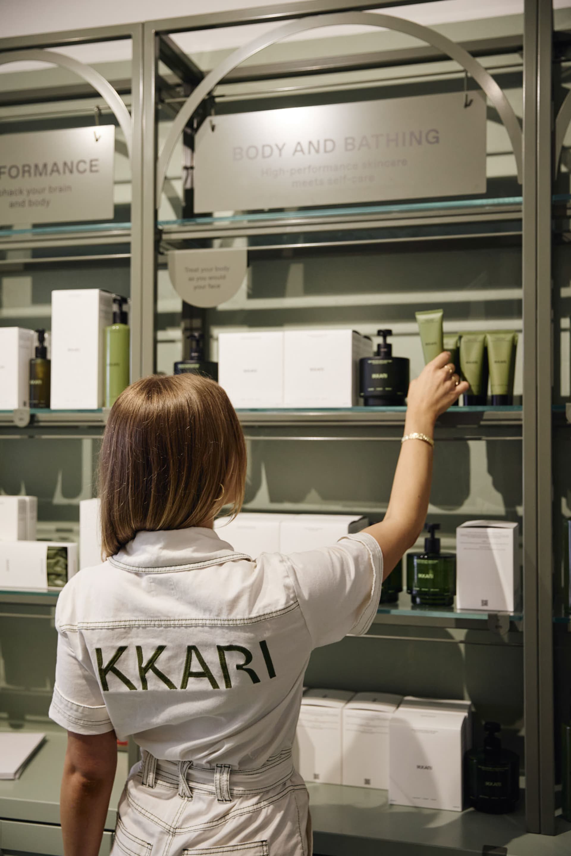 A woman in a white shirt is arranging bottles on a glass shelf in a retail setting.
