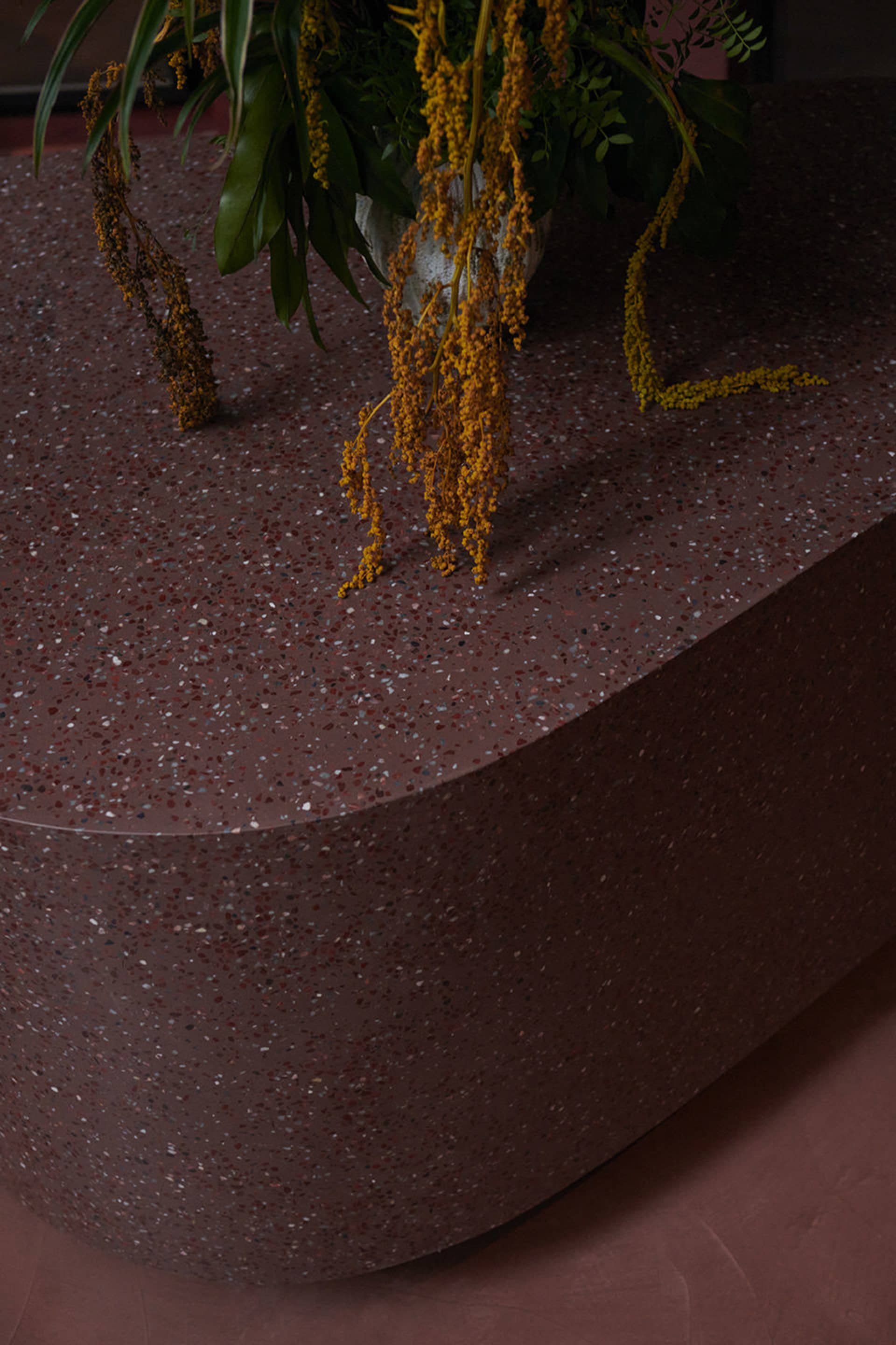 A sleek, modern table made of red terrazzo with a smooth, rounded surface, featuring a plant with yellow flowers in a white v