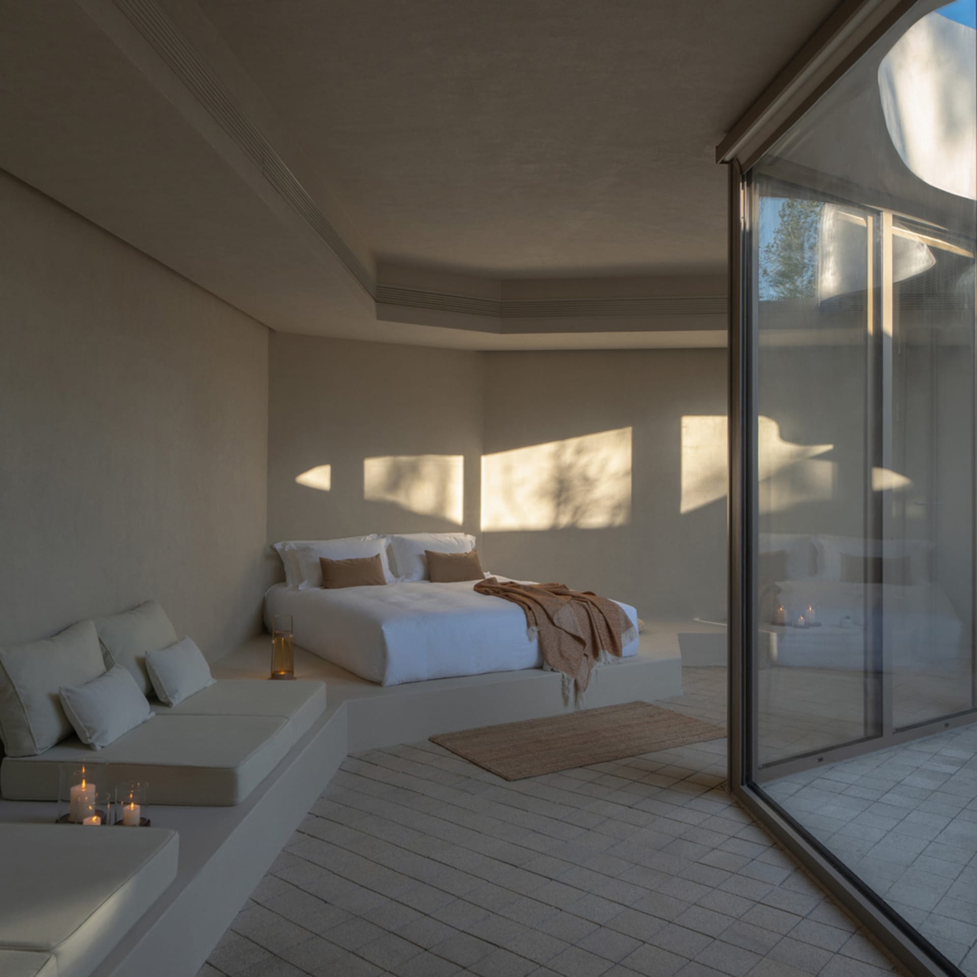 A minimalist bedroom featuring a concrete and glass platform bed, with white bedding and a window allowing sunlight to stream