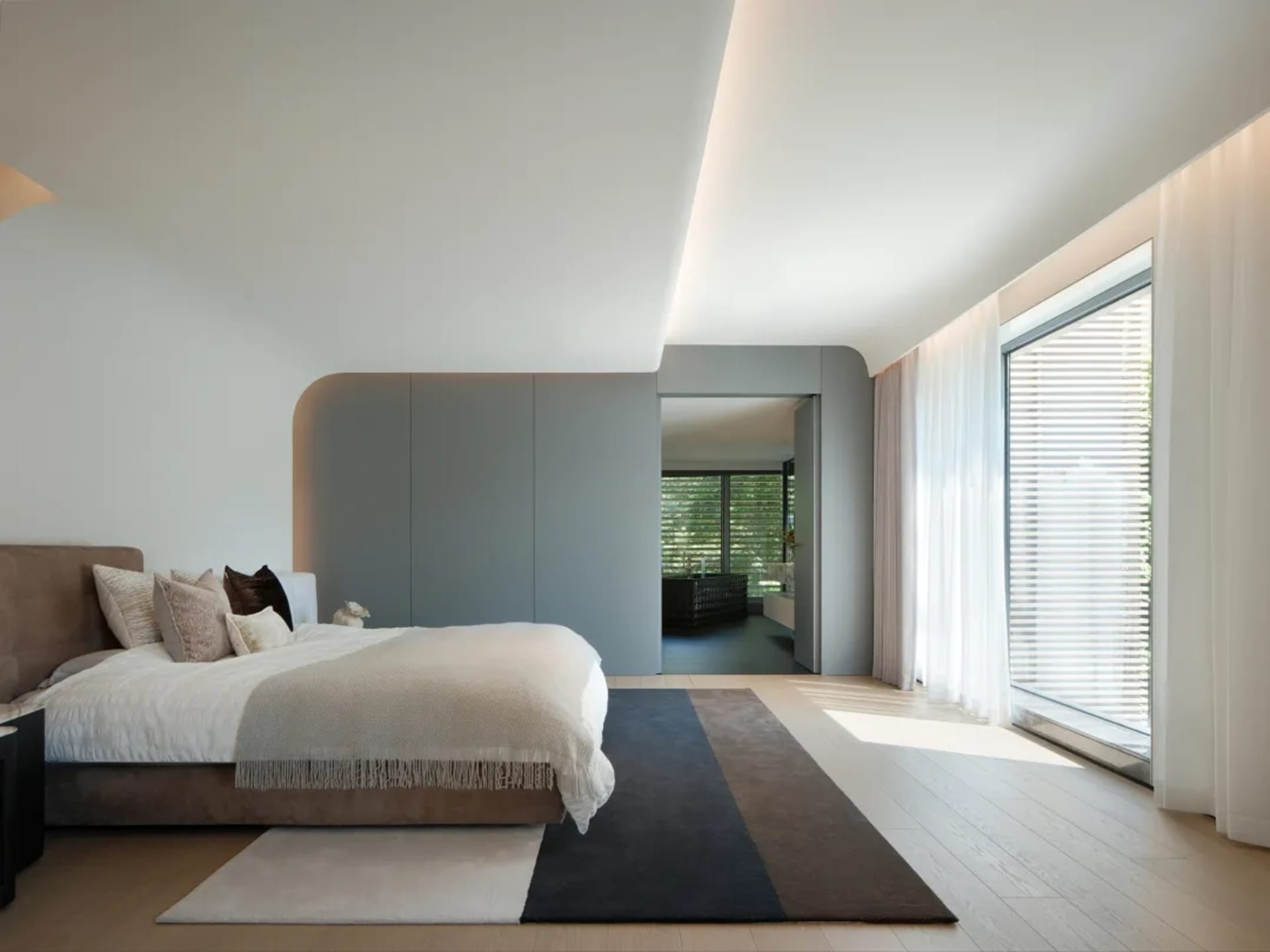 A minimalist bedroom with a bed, pillows, and a rug on a wooden floor, featuring curved architectural elements and large wind