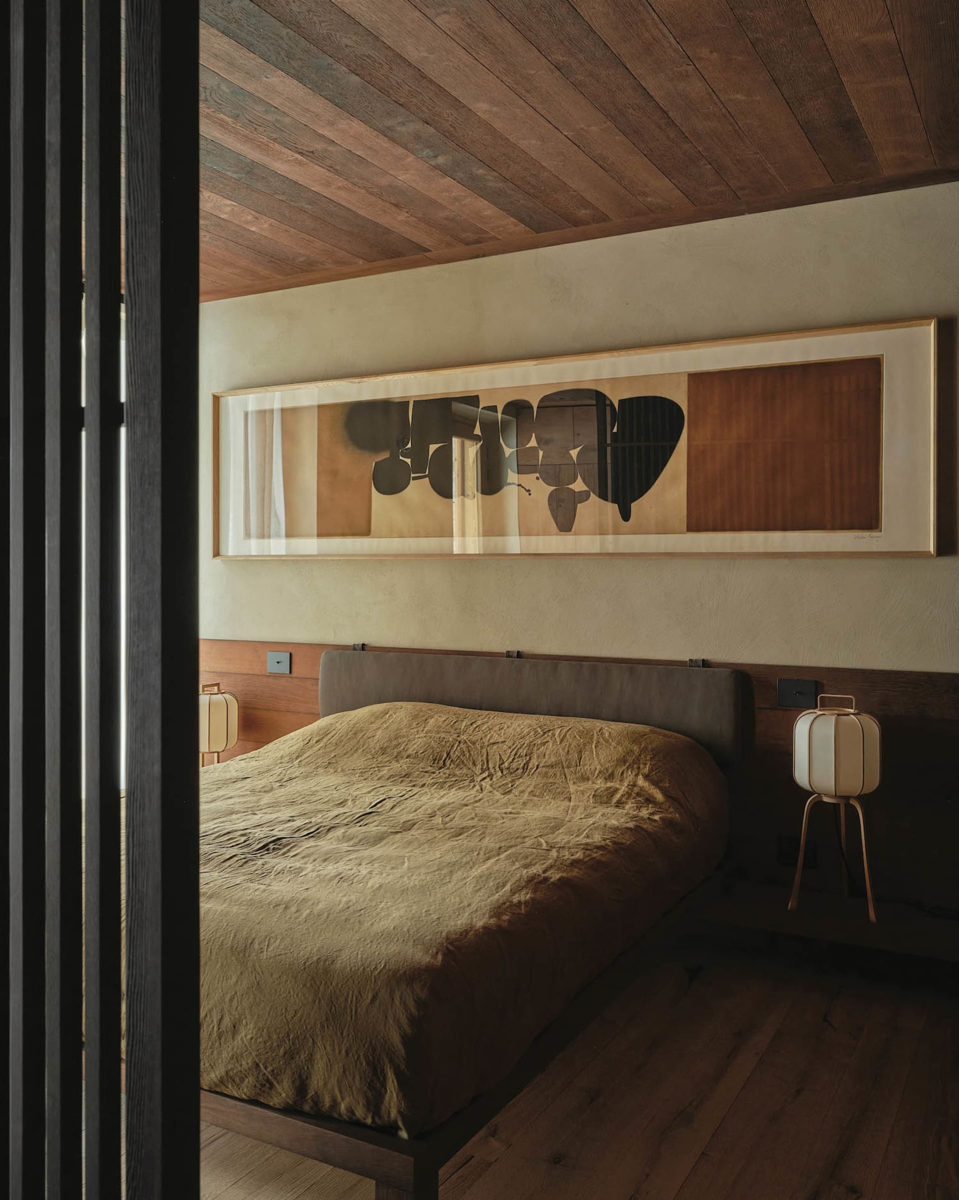 A minimalist bedroom featuring a wooden bed frame and headboard, with a large framed abstract painting above it.