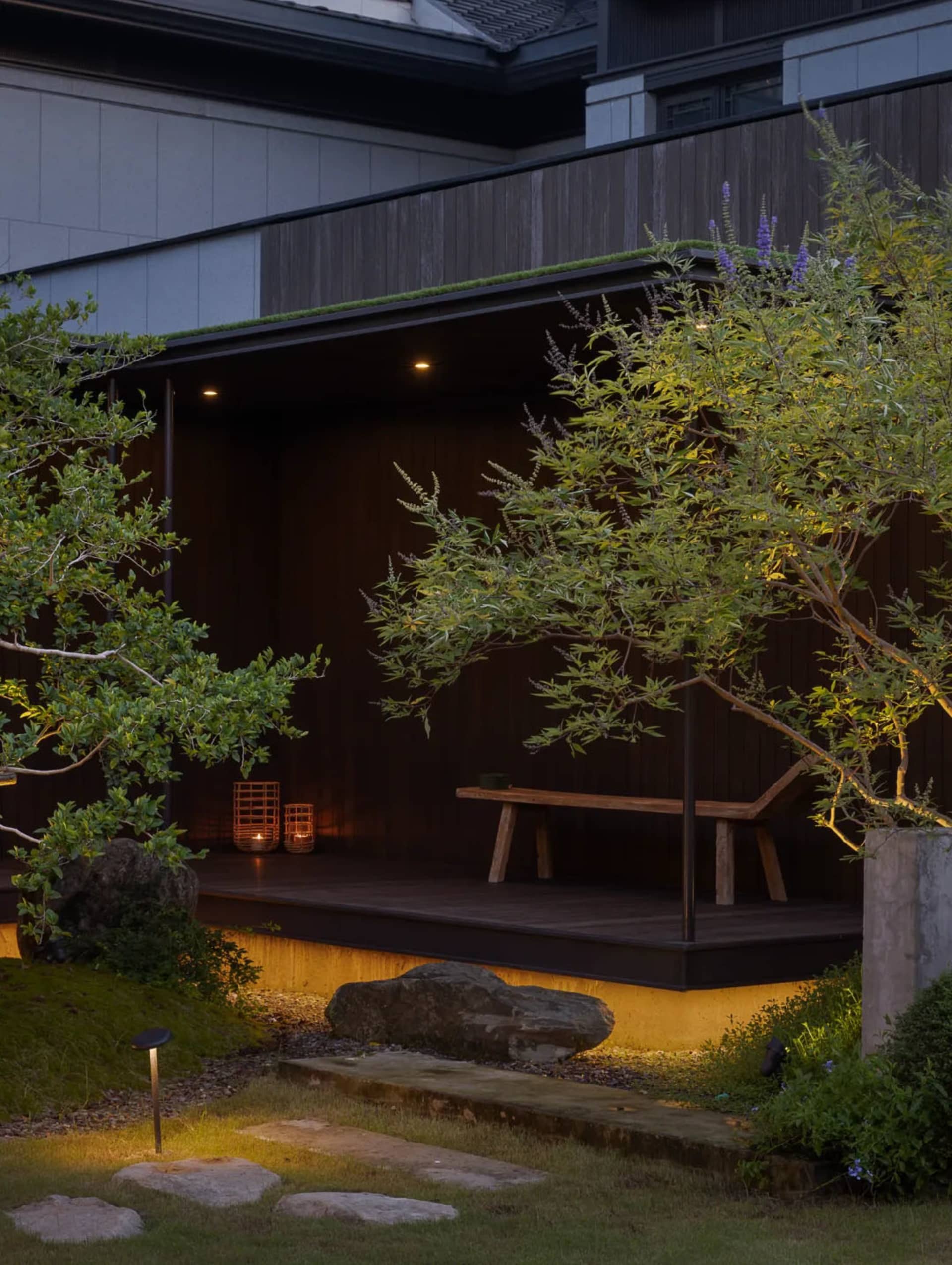 A minimalist garden featuring a wooden platform, a stone bench, and rock formations illuminated by warm LED lights.