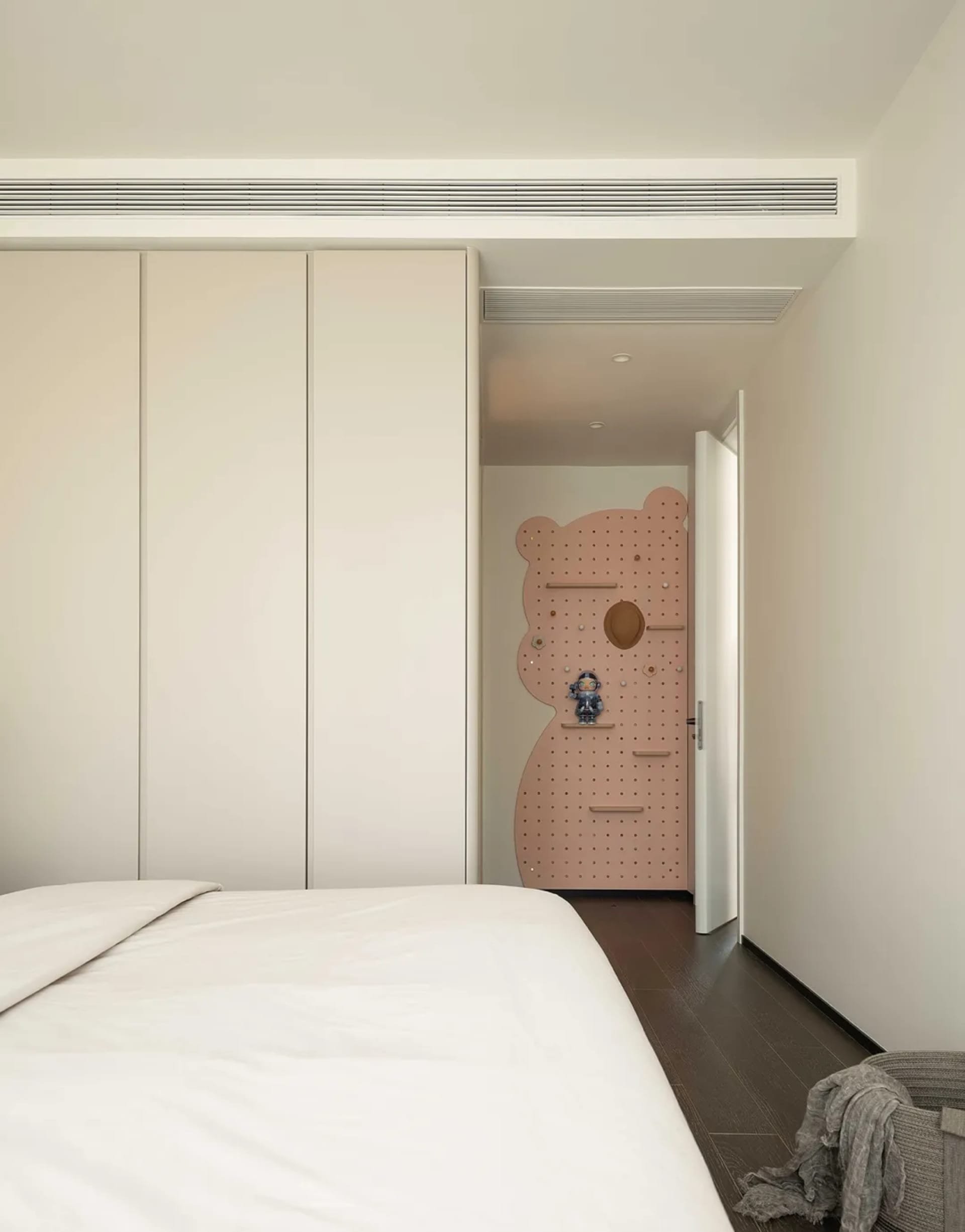 A minimalist bedroom featuring a white bed with a modern design, surrounded by white closet doors and a pink pegboard wall fe