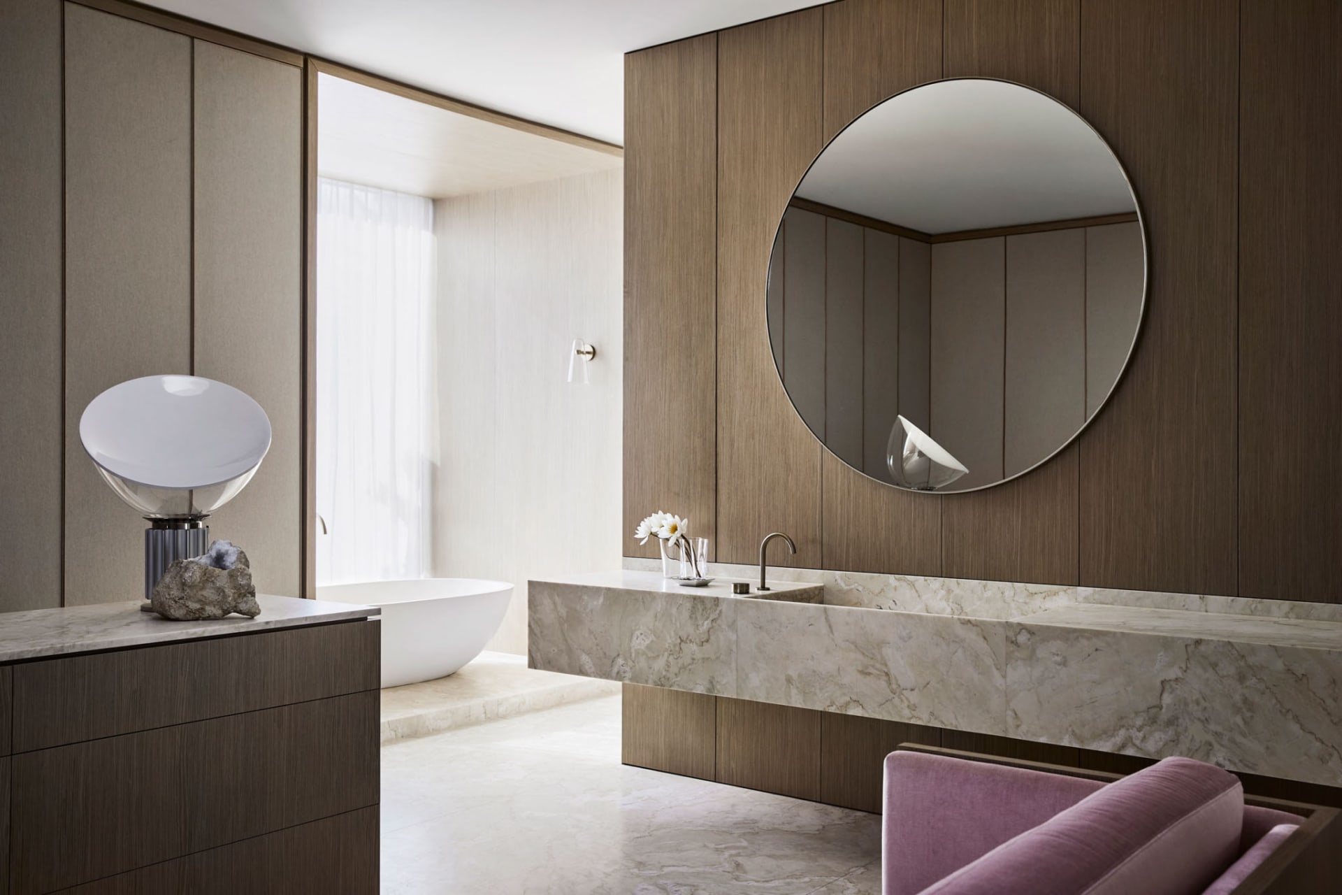 A modern bathroom with a large round mirror, marble sink, wooden panels, and a freestanding bathtub.