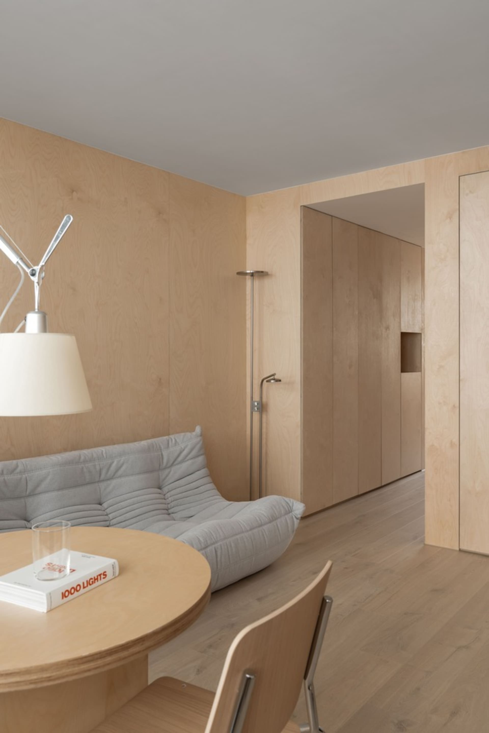 A minimalist living room featuring a grey sofa, round wooden table, and wood-paneled walls.