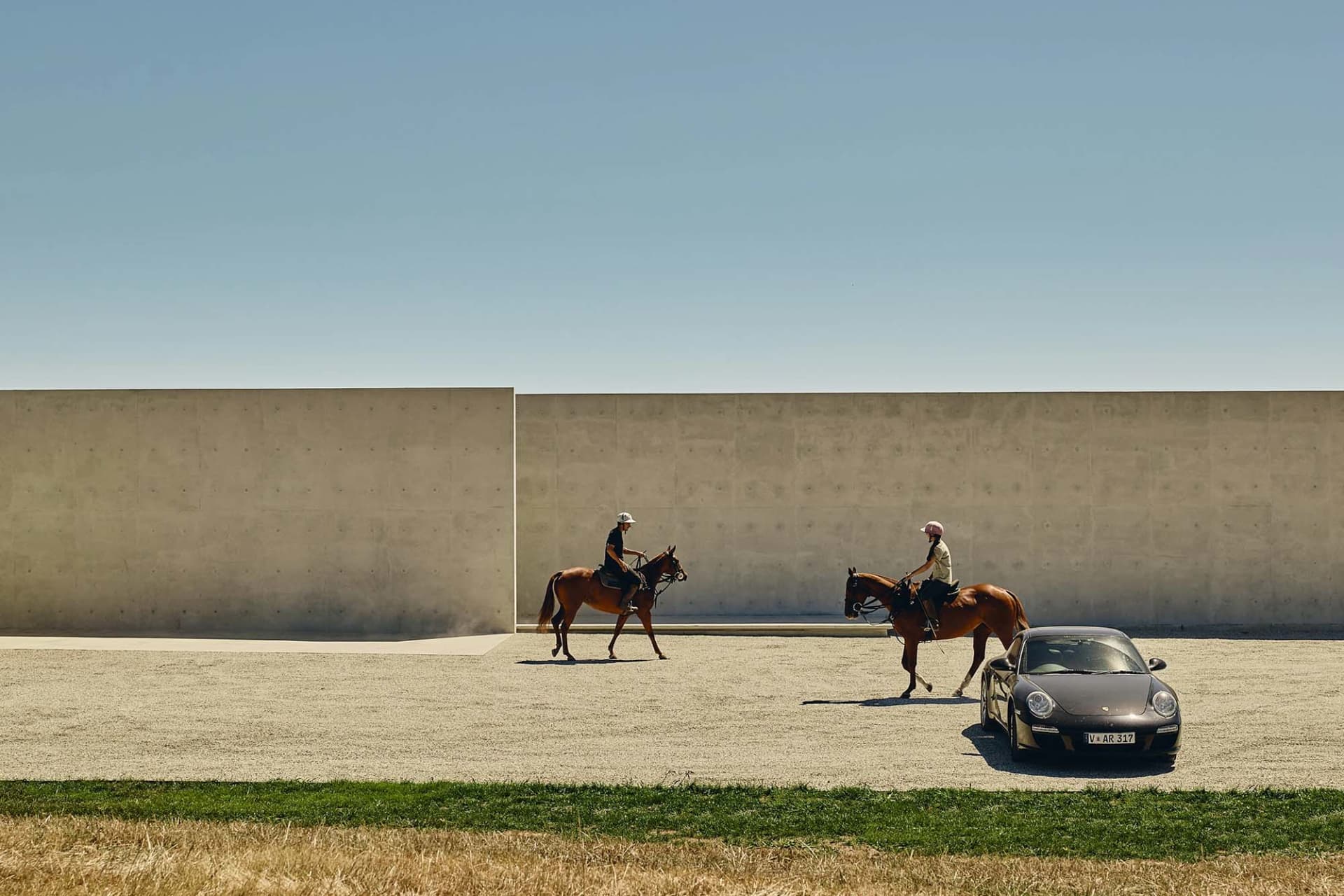 Two people riding horses in front of a concrete wall, with a car parked nearby.
