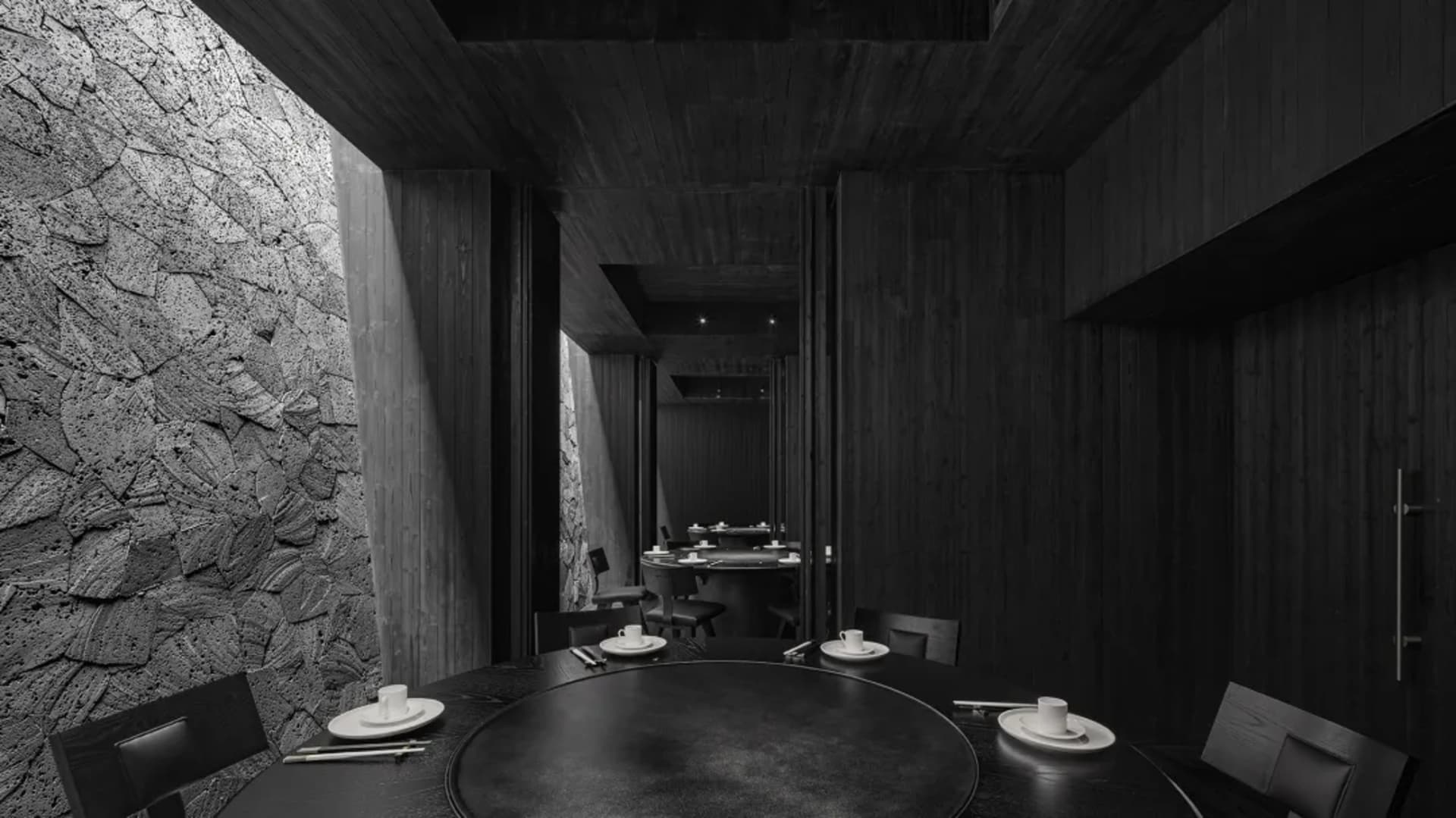 A modern restaurant interior with black tableware, chairs, and a stone wall, featuring a round table and minimalist decor.