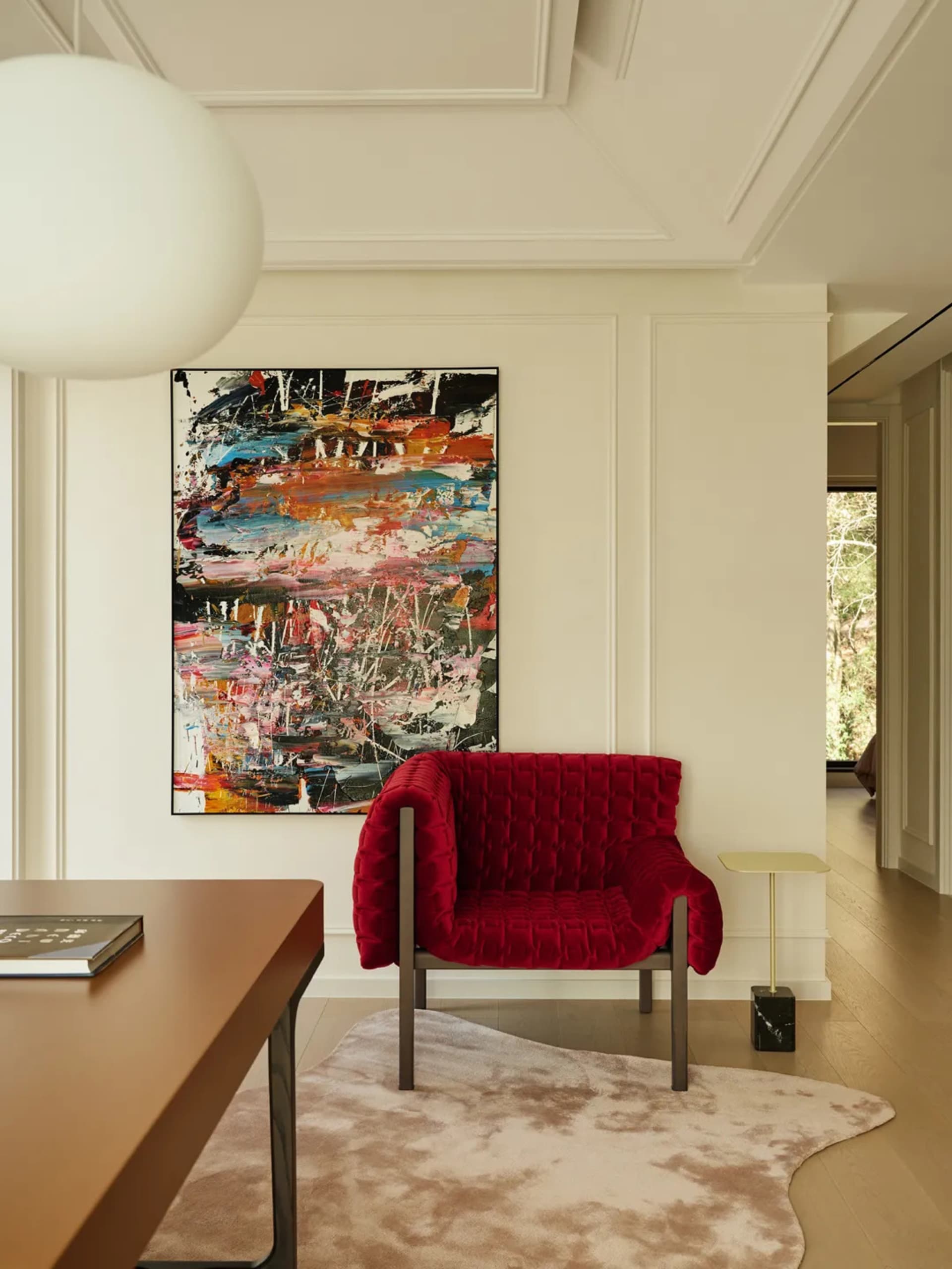 A modern living room with a red velvet armchair on a cowhide rug, framed artwork on the wall, and a wooden desk with a book.