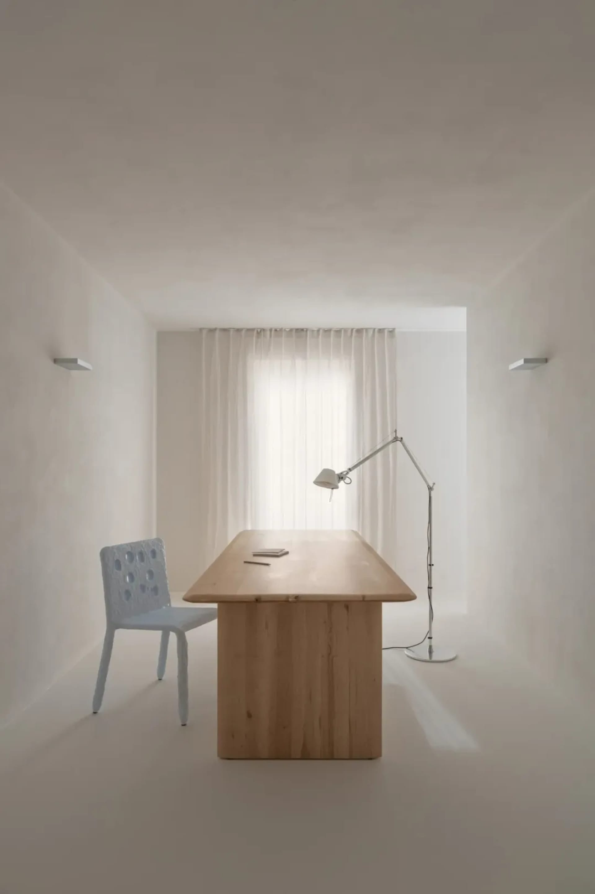 A minimalist office space featuring a wooden desk and chair next to a window with sheer curtains, illuminated by a lamp.