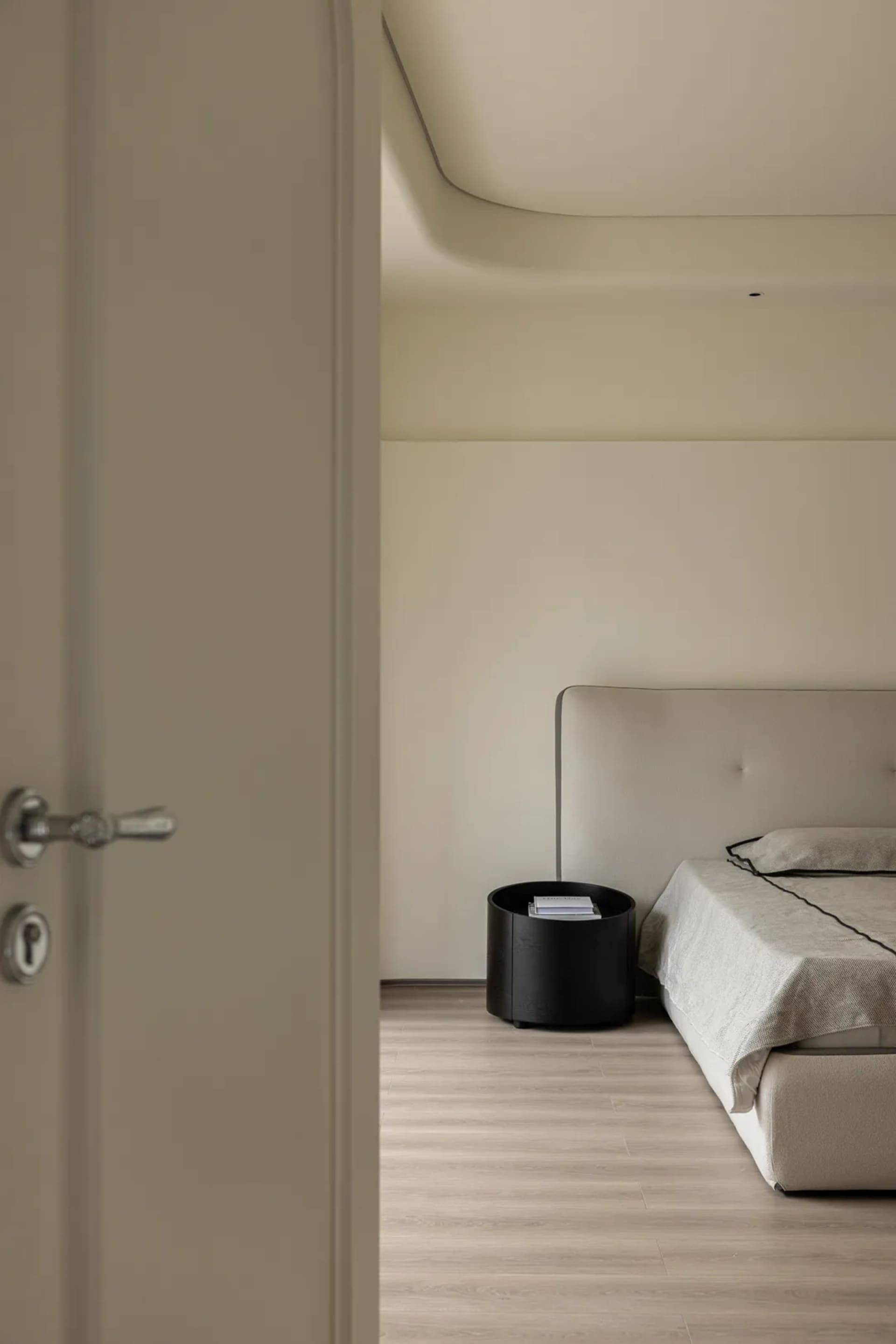 A minimalist bedroom with a grey upholstered bed, black side table, and wooden flooring.