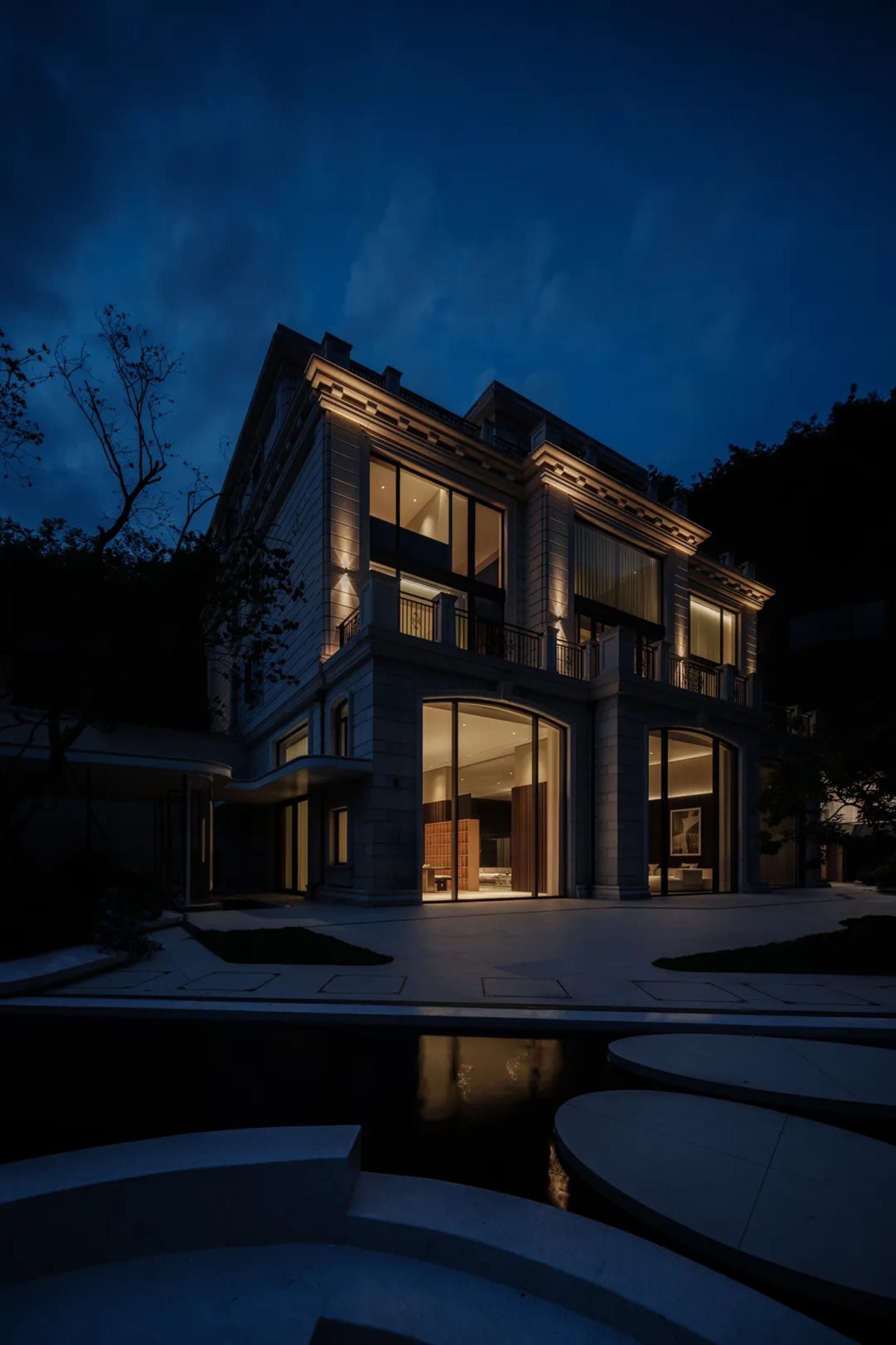 一座現代住宅建築採用混凝土外觀,夜間窗戶燈光亮起,庭院中設有水景