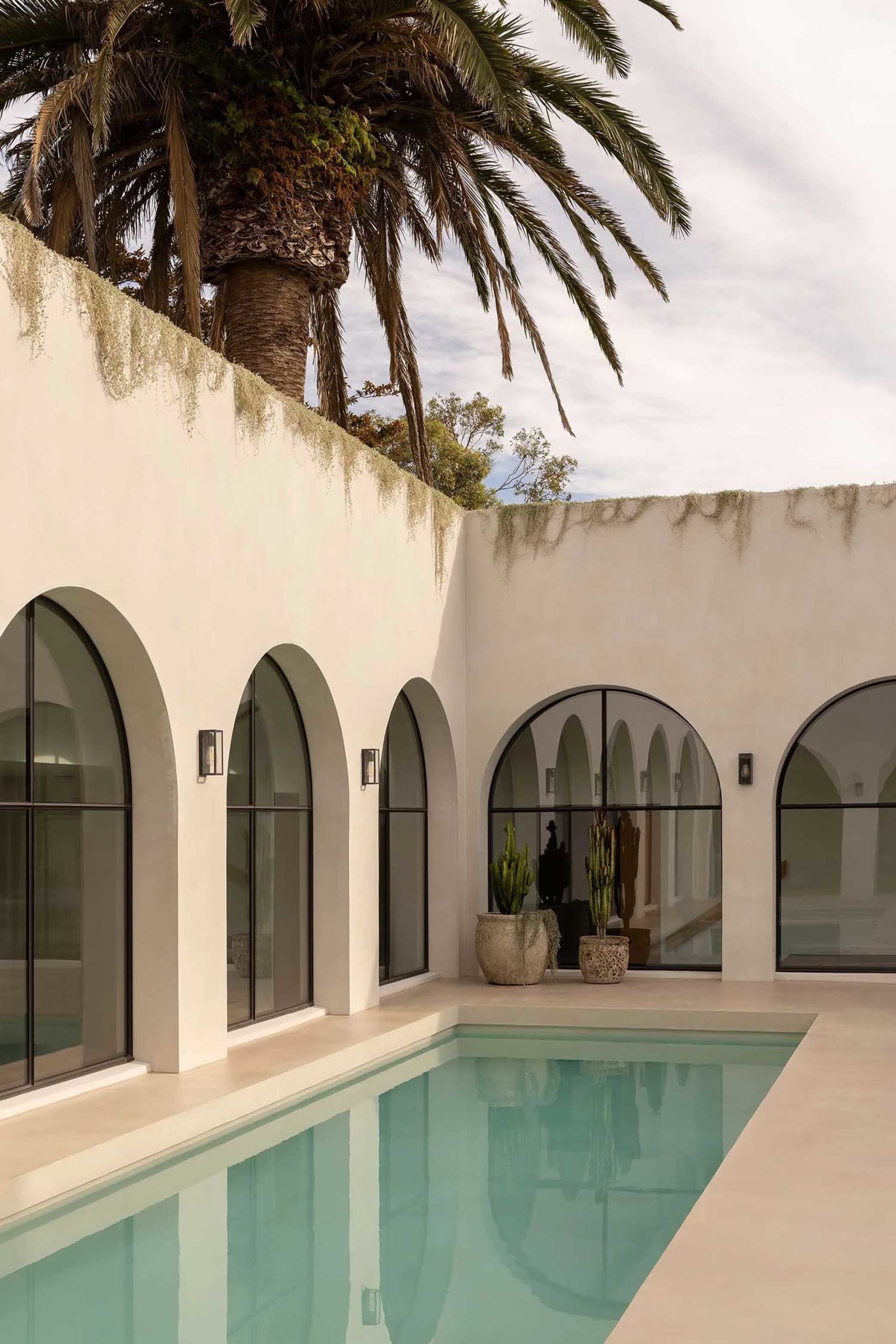 Modern house with white plaster exterior, arched windows, and a swimming pool surrounded by palm trees.