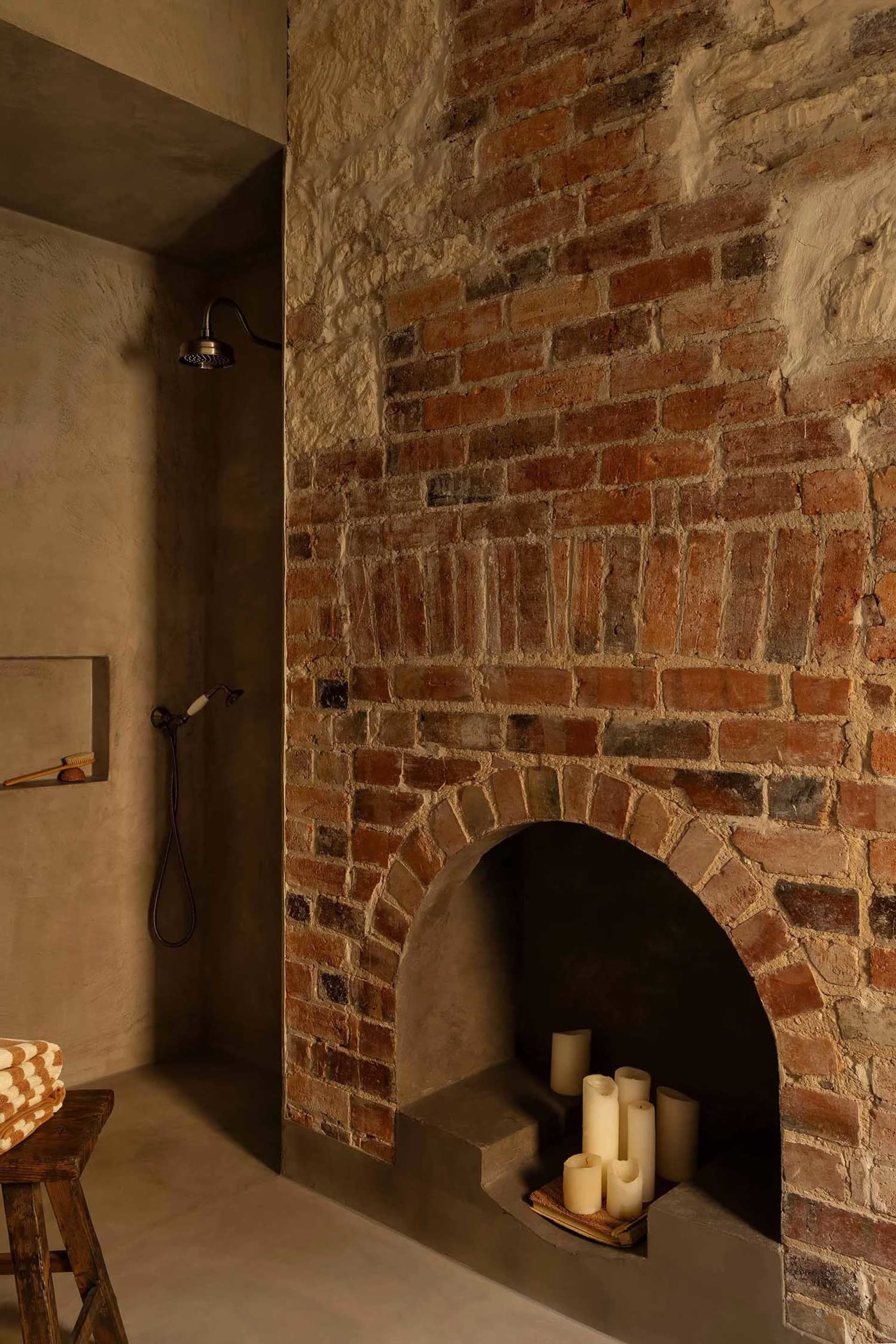 A minimalist bathroom showcases a shower, candles, and a wooden stool in a space with concrete walls and an exposed brick fir