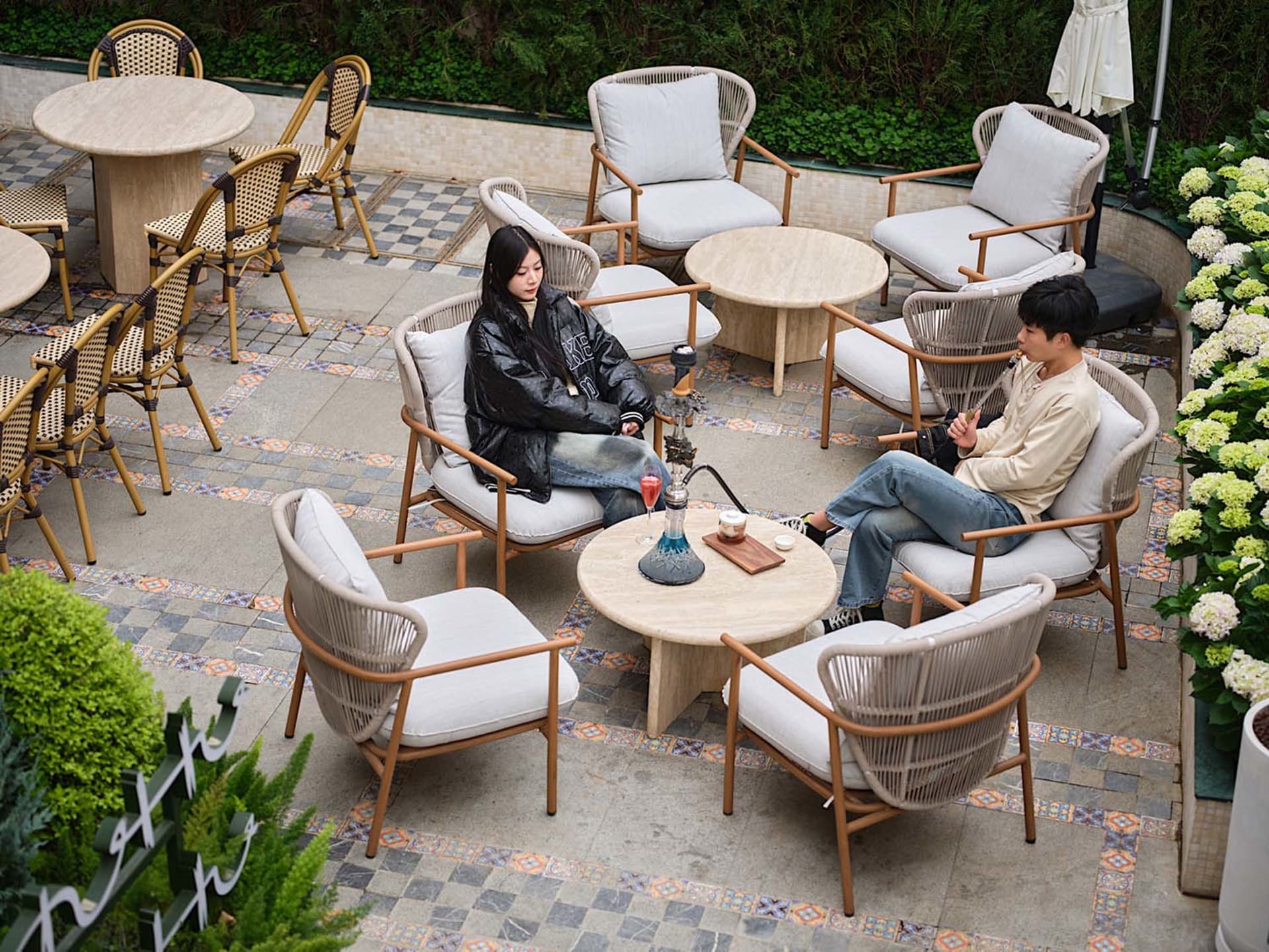 Two individuals sitting on wicker patio furniture with white cushions, talking at a round table adorned with a hookah and dri
