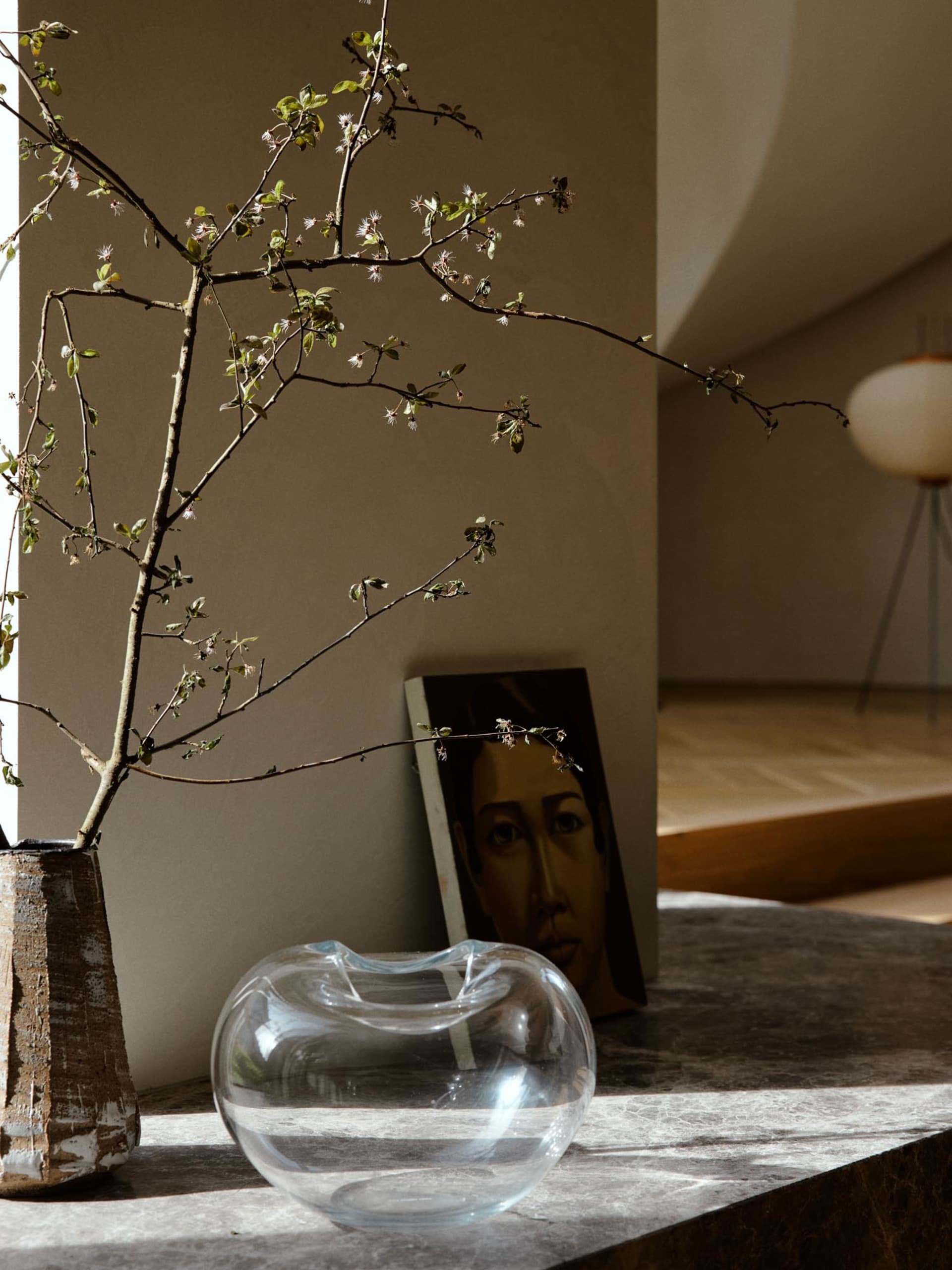 A minimalist interior scene featuring a glass vase holding a branch with small flowers and leaves, placed on a stone counter