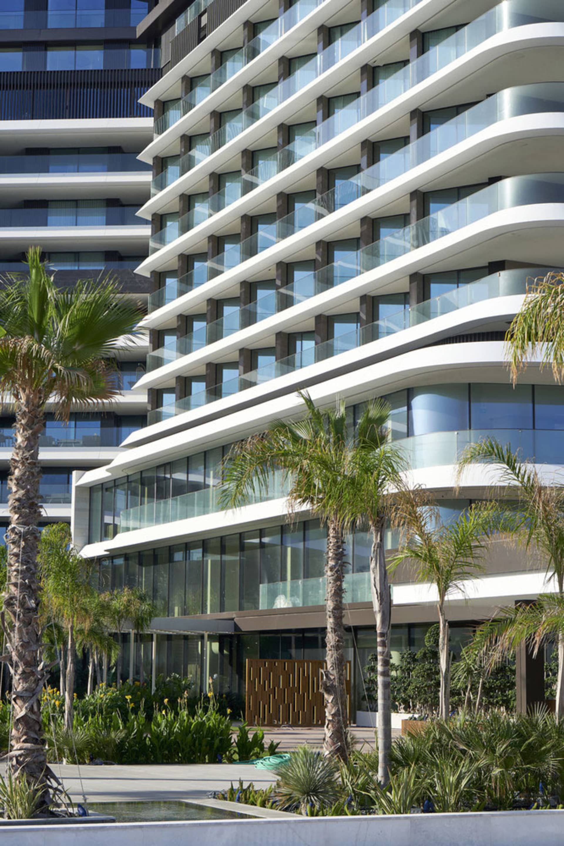 Modern residential building with glass windows and concrete balconies, surrounded by palm trees and greenery.