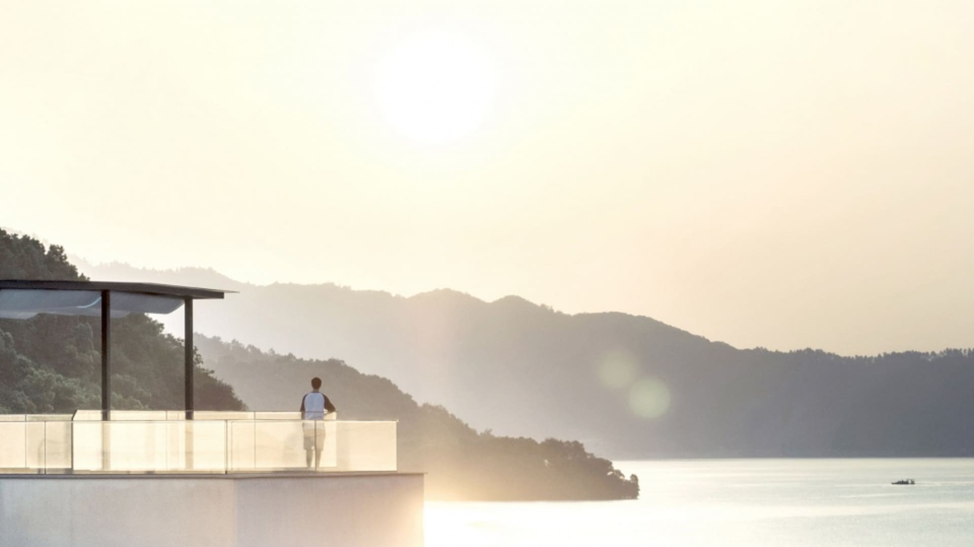 Person standing on a balcony overlooking a serene lake surrounded by mountains during sunset.