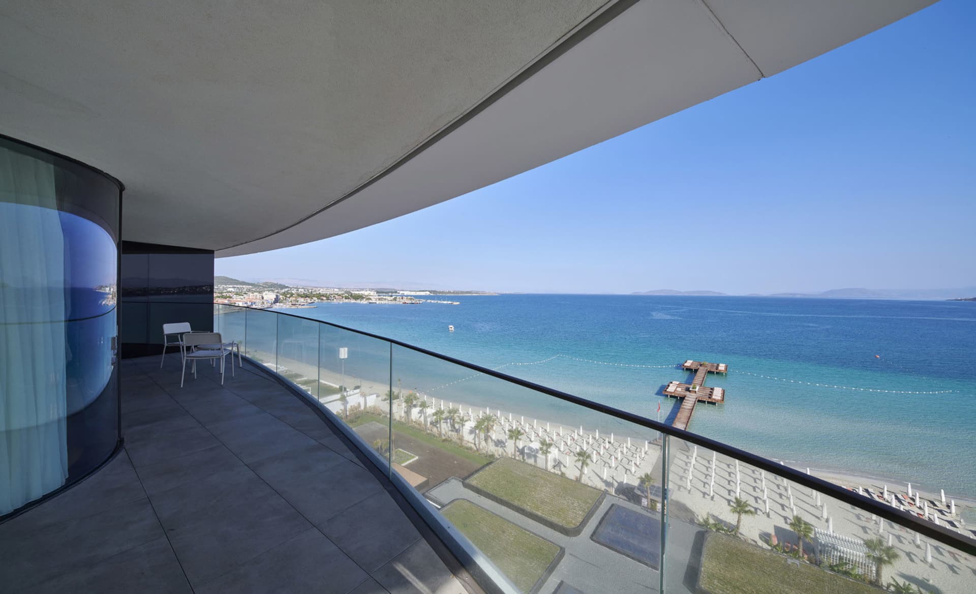 Modern, minimalist apartment balcony overlooking the ocean with clear blue skies and a clear view of the beach.
