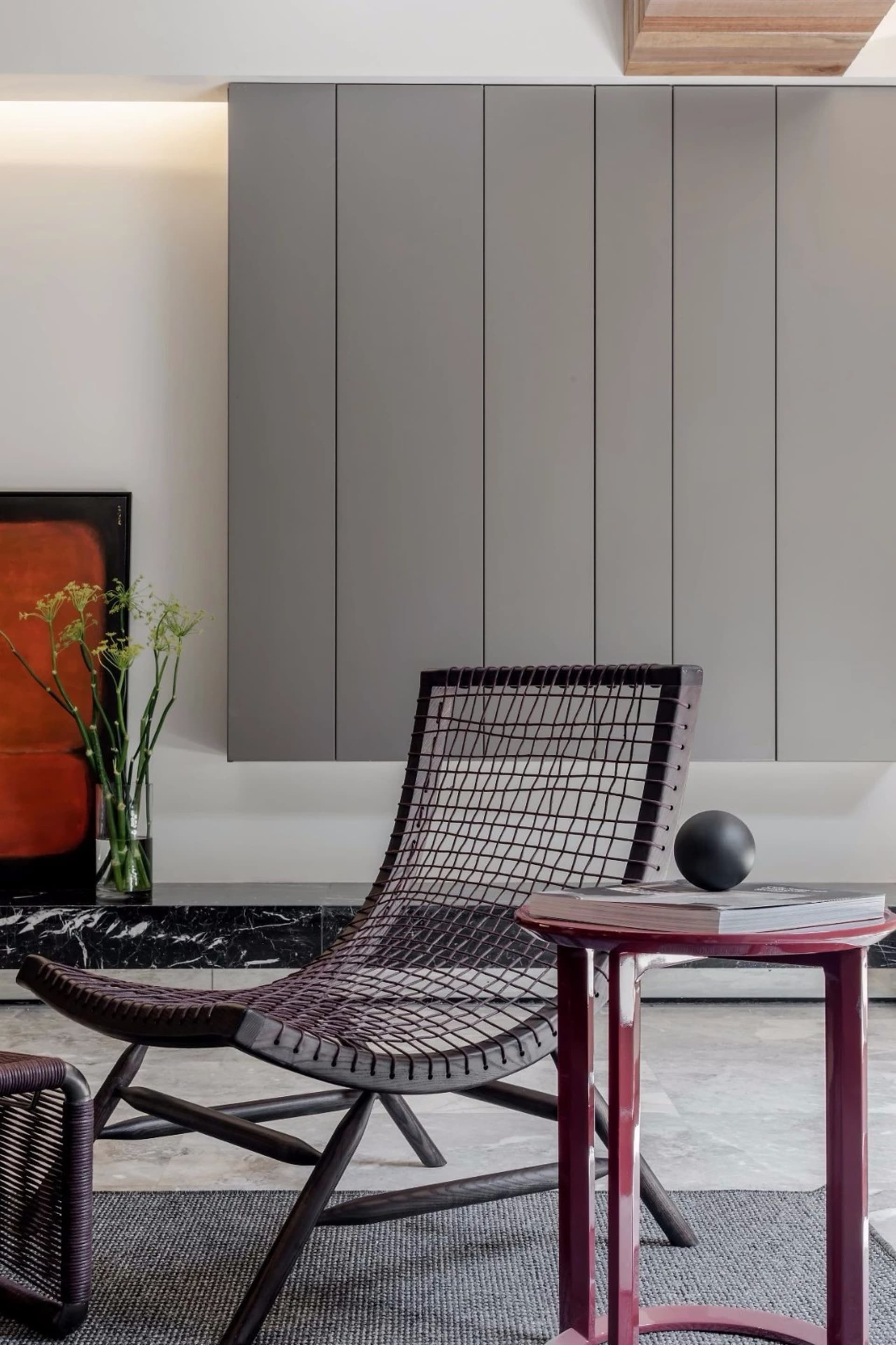 A minimalist living room with a black metal-net chair, a red side table with a black ball and books, and a black marble count
