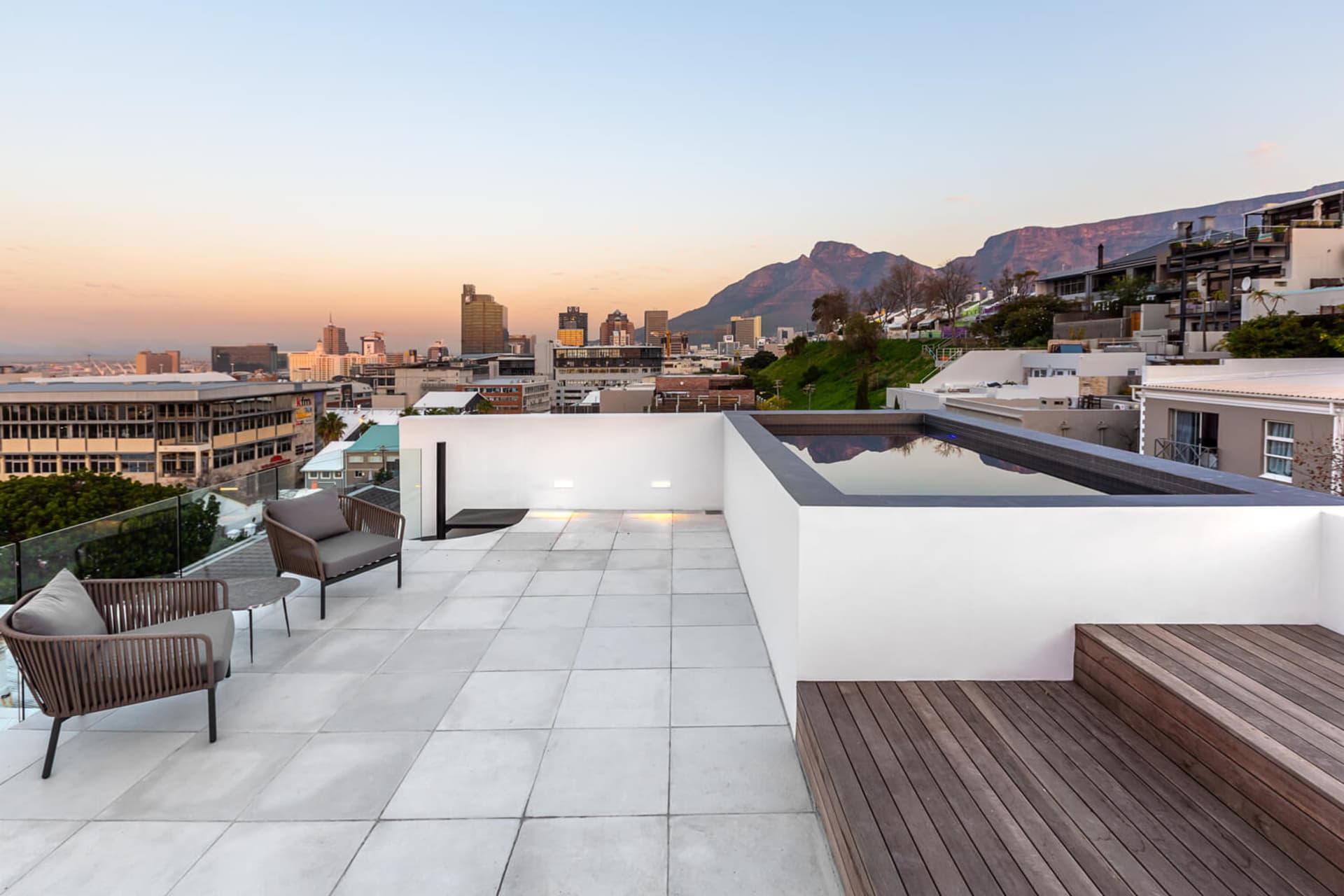 Modern rooftop deck with white concrete tiles, minimal furniture, and a view of the city skyline and mountains at sunset.