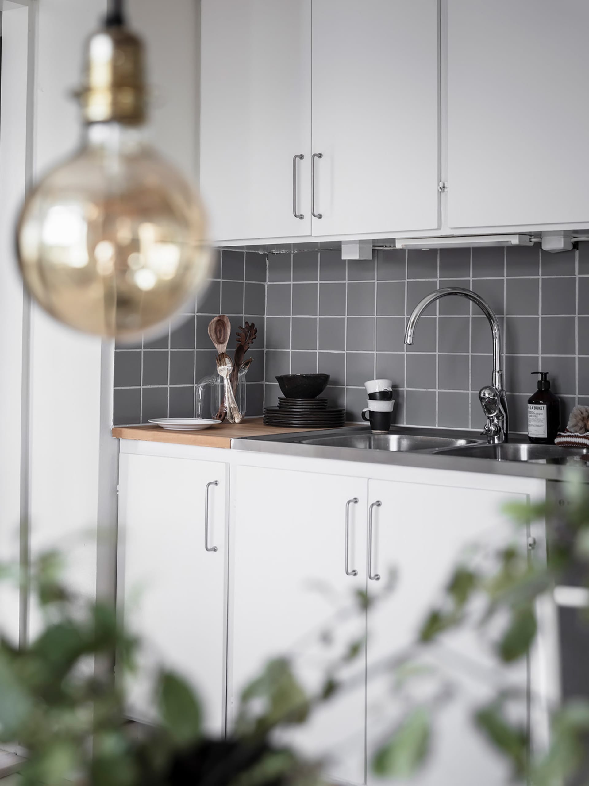 A modern, minimalist kitchen with white cabinets, a stainless steel sink, gray tiled backsplash, and a hanging light bulb.