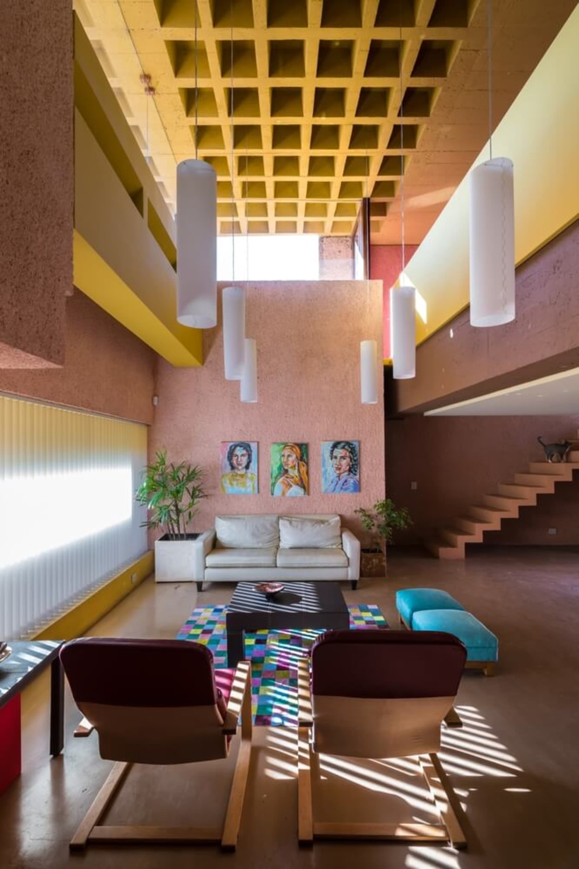 A modern living room with concrete walls and floors, featuring a white sofa, colorful rugs, hanging lamps, and a staircase in