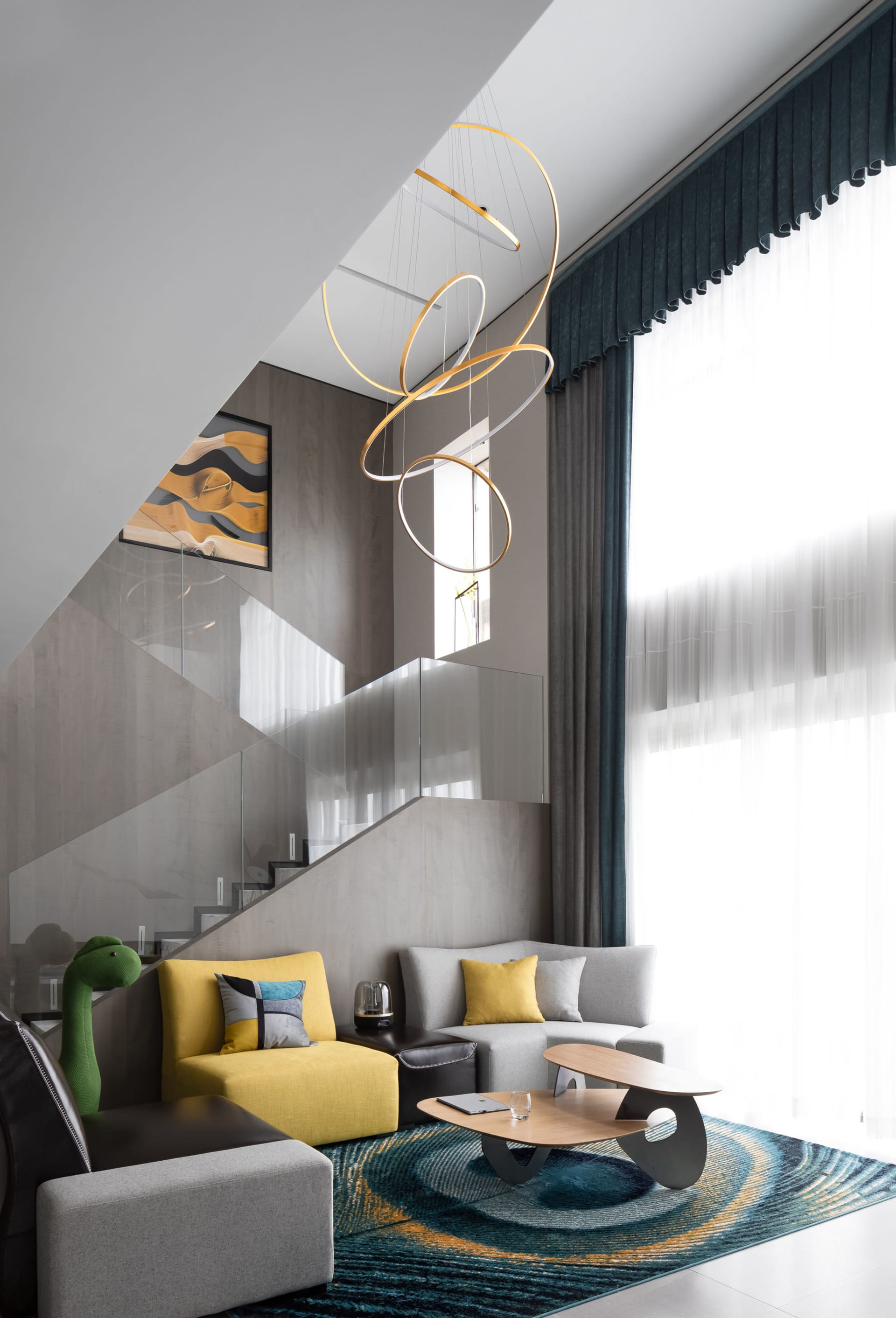 Modern living room with yellow couch, grey sofa, glass staircase, and abstract lighting fixture