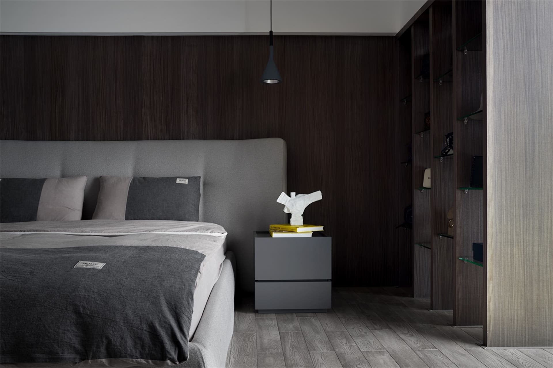 A modern minimalist bedroom featuring wood-paneled walls, a grey upholstered bed, and a grey wooden nightstand with books and