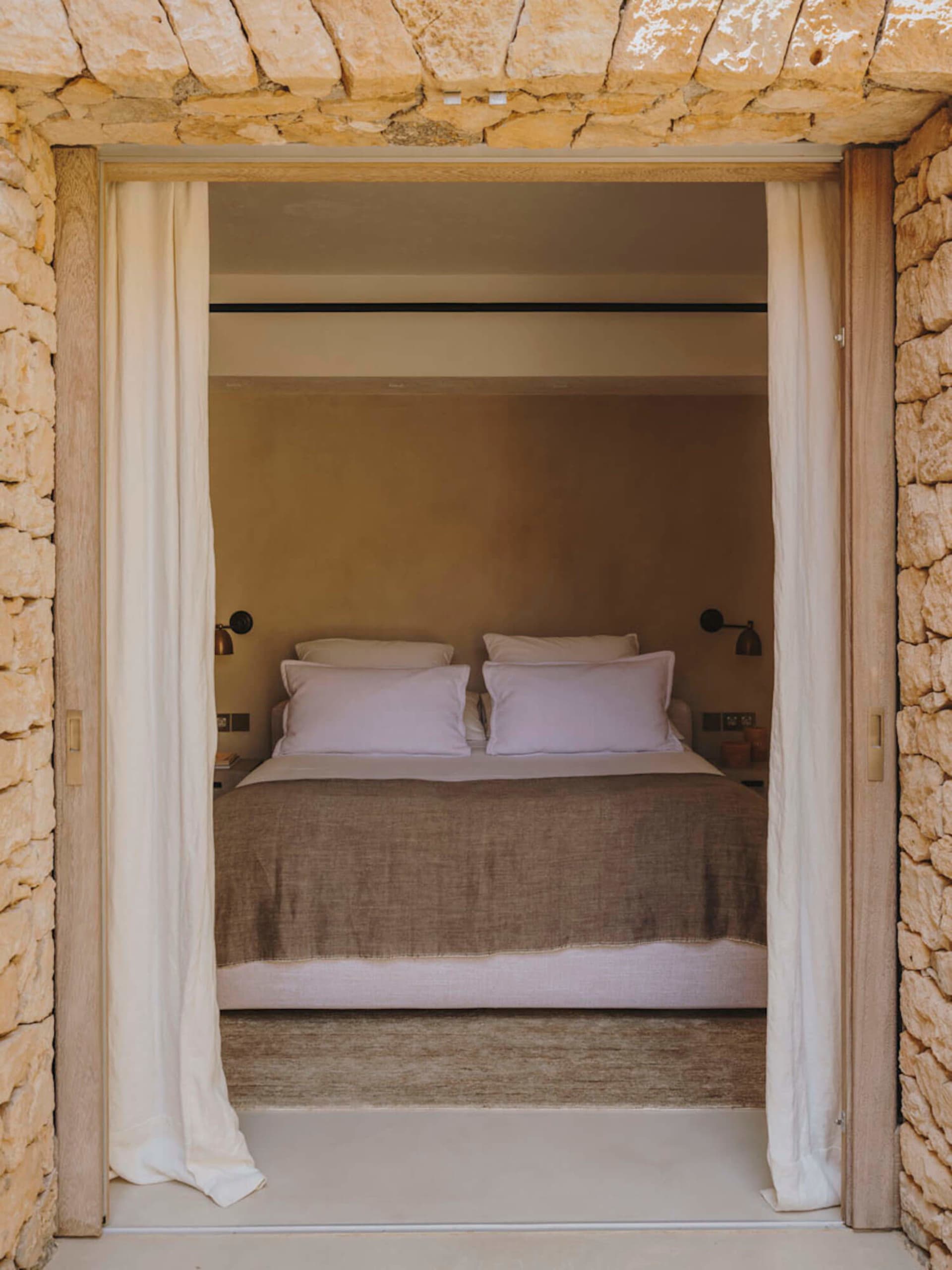 A minimalist bedroom with a bed covered in lavender linen sheets and a stone wall, enclosed by white curtains.