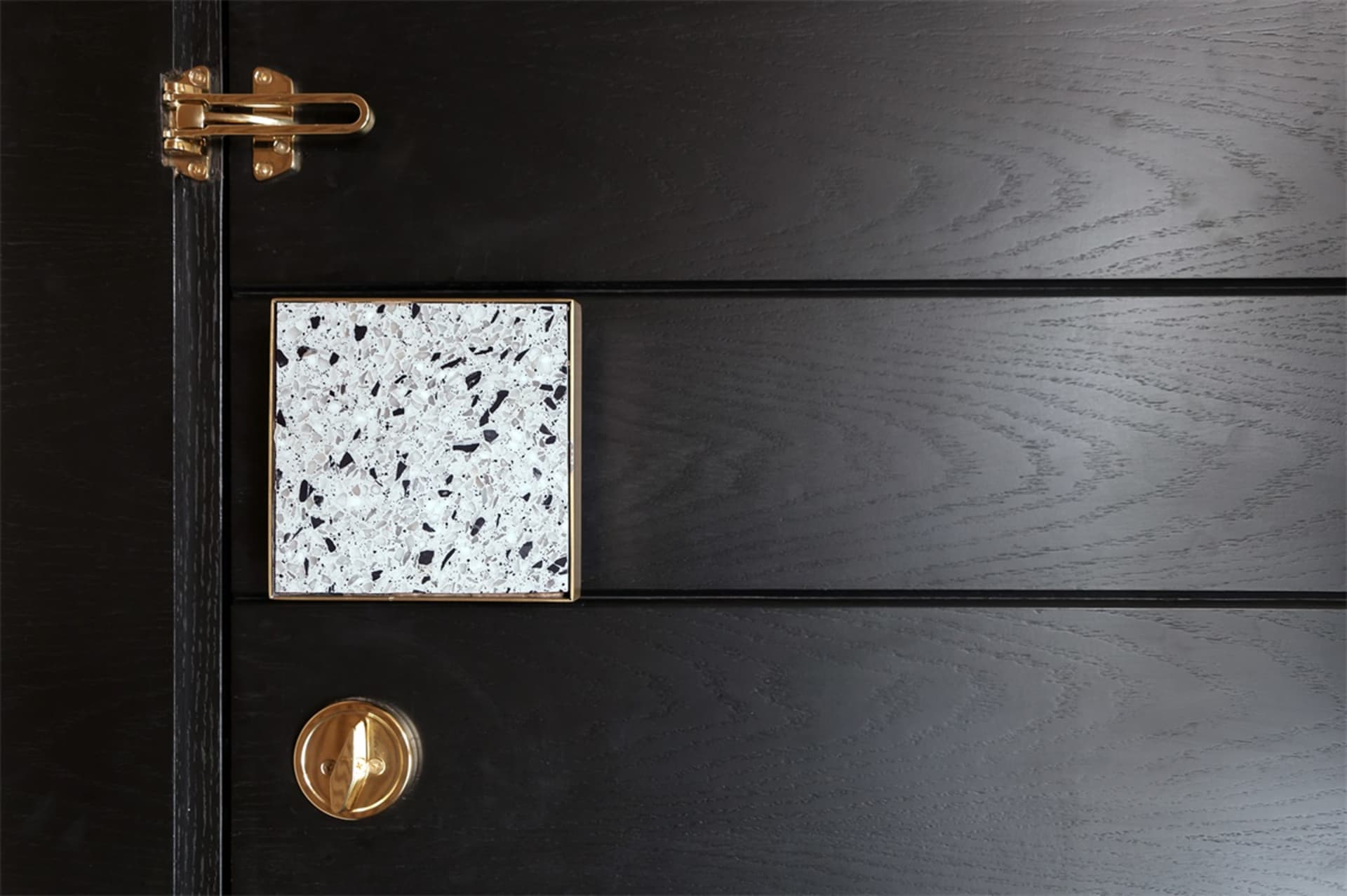 An elegant, black-colored wooden door with a geometric, white speckled terrazzo tile and a gold latch.