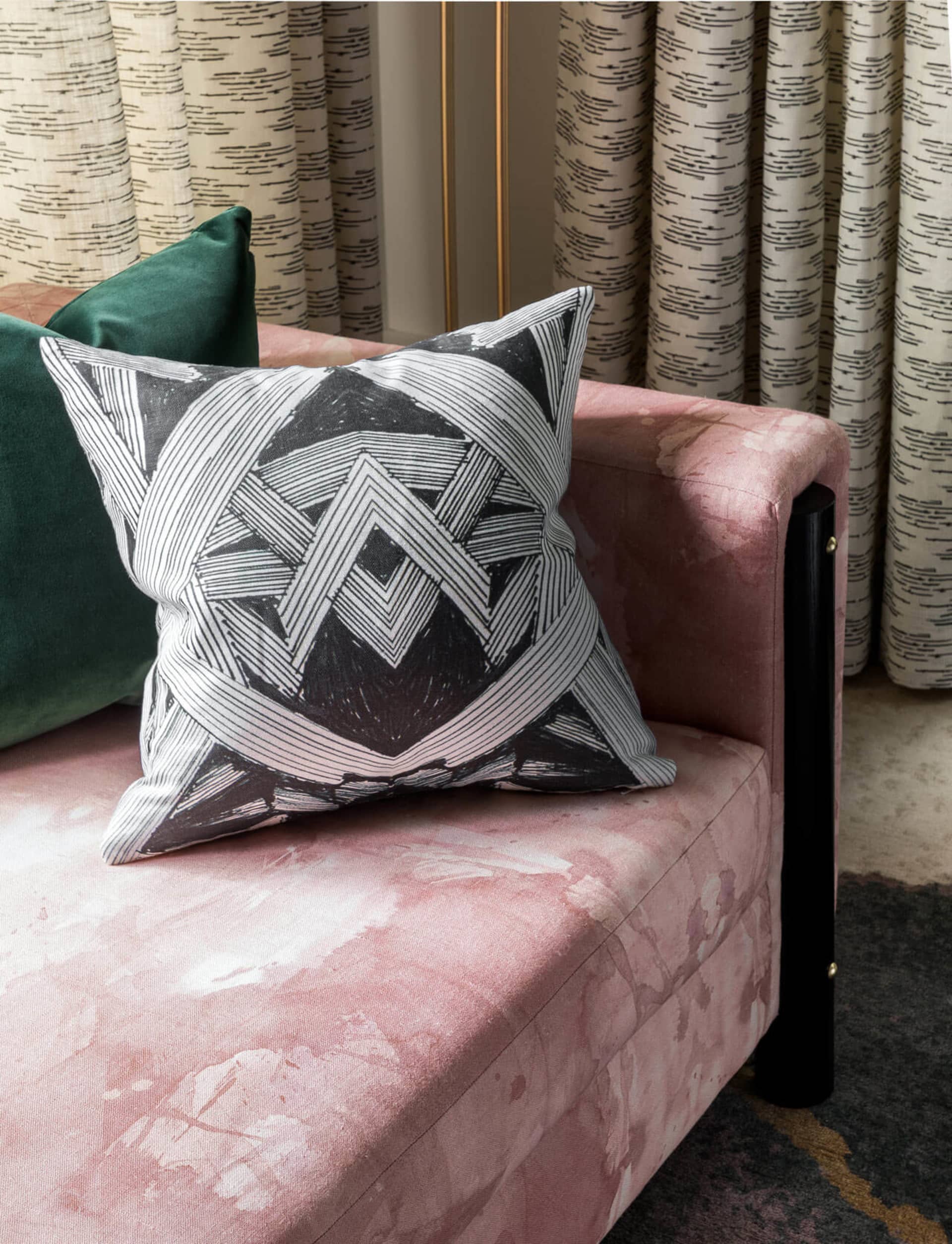 A modern couch with a pink velvet upholstery and patterned pillows against a backdrop of striped curtains and a patterned flo