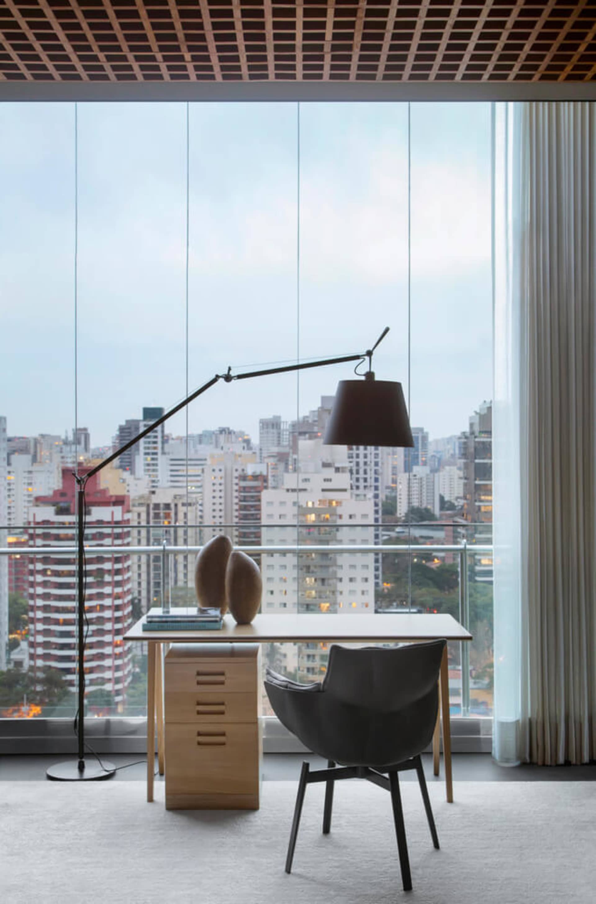 A minimalist office with a desk, chair, and floor lamp in front of a large window with a cityscape view.