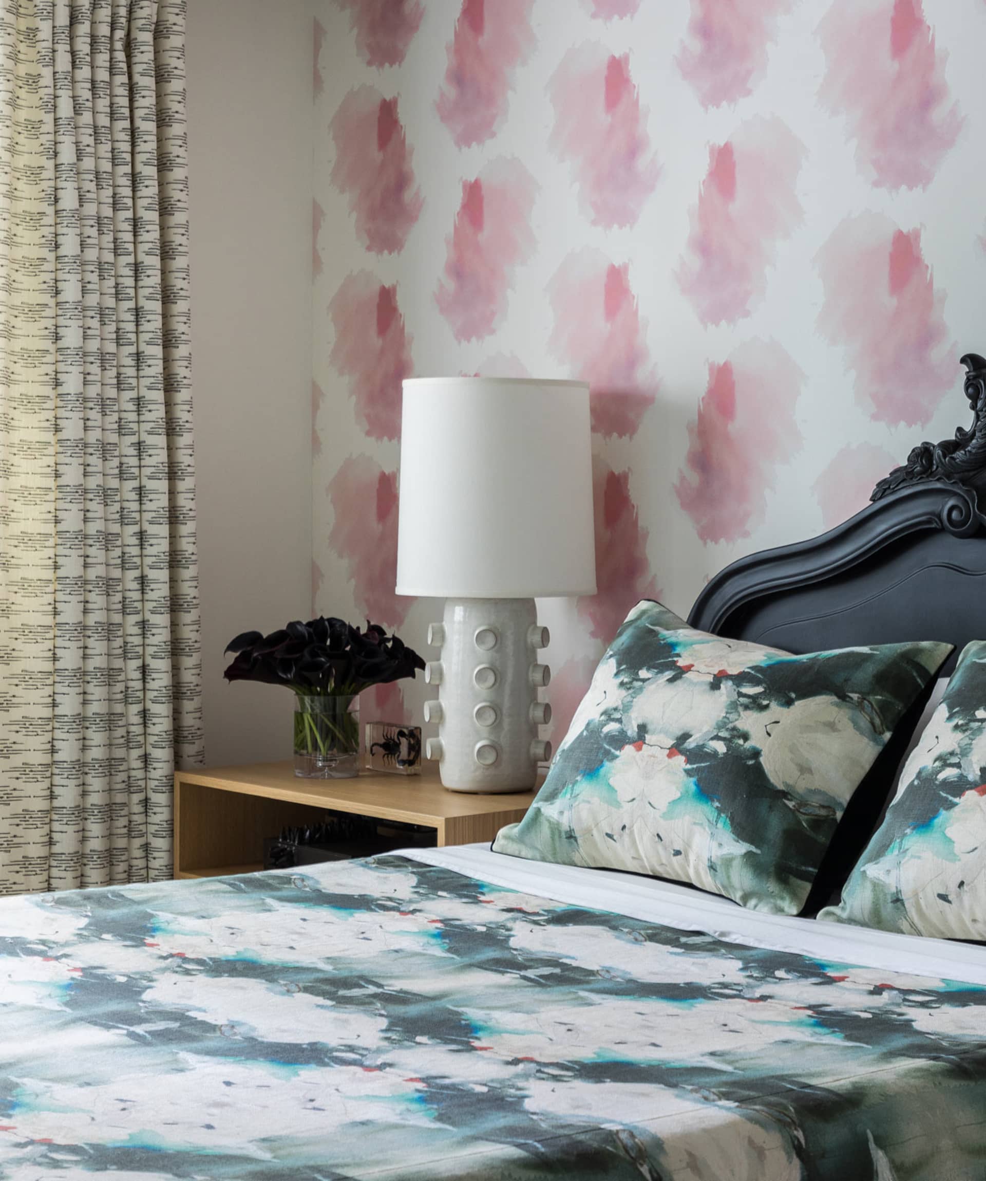 Modern bedroom with a patterned bedspread, floral pillows, and a white lamp on a wooden shelf.