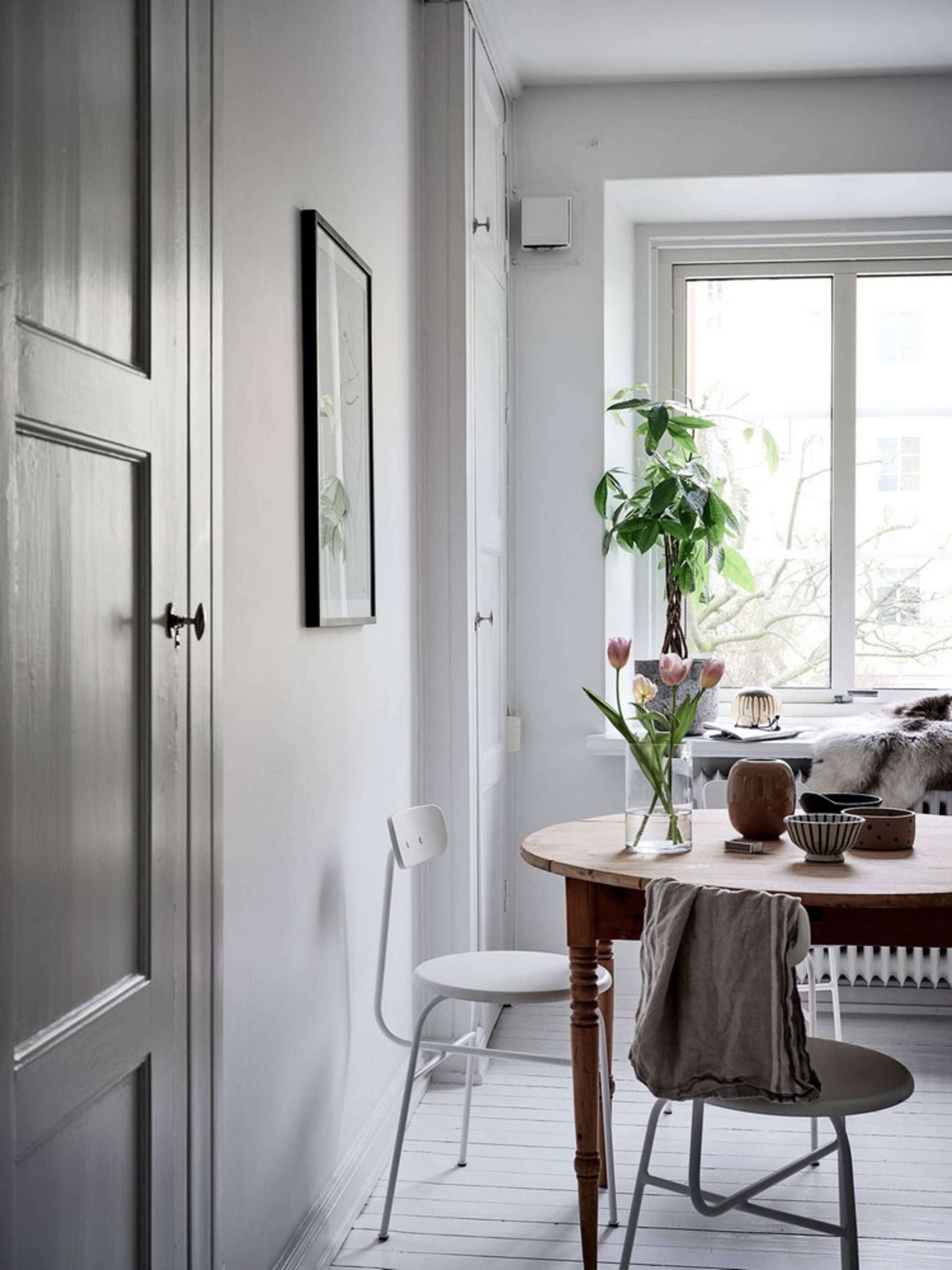 Modern, white minimalist interior with wood dining table and chairs, minimalist art, and a plant on the window sill.