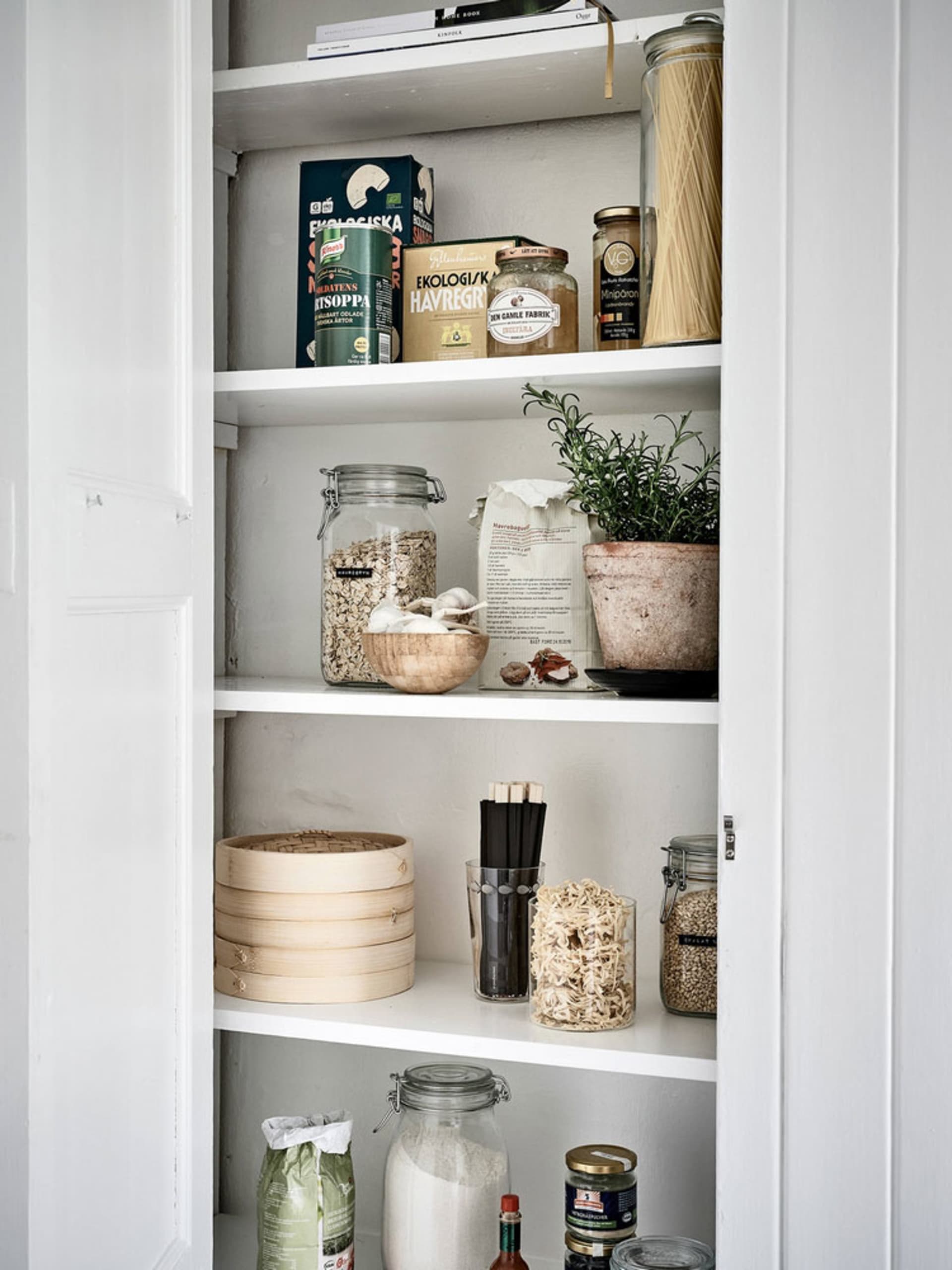 A white kitchen cupboard with organized shelves containing various food items, utensils, and plants.