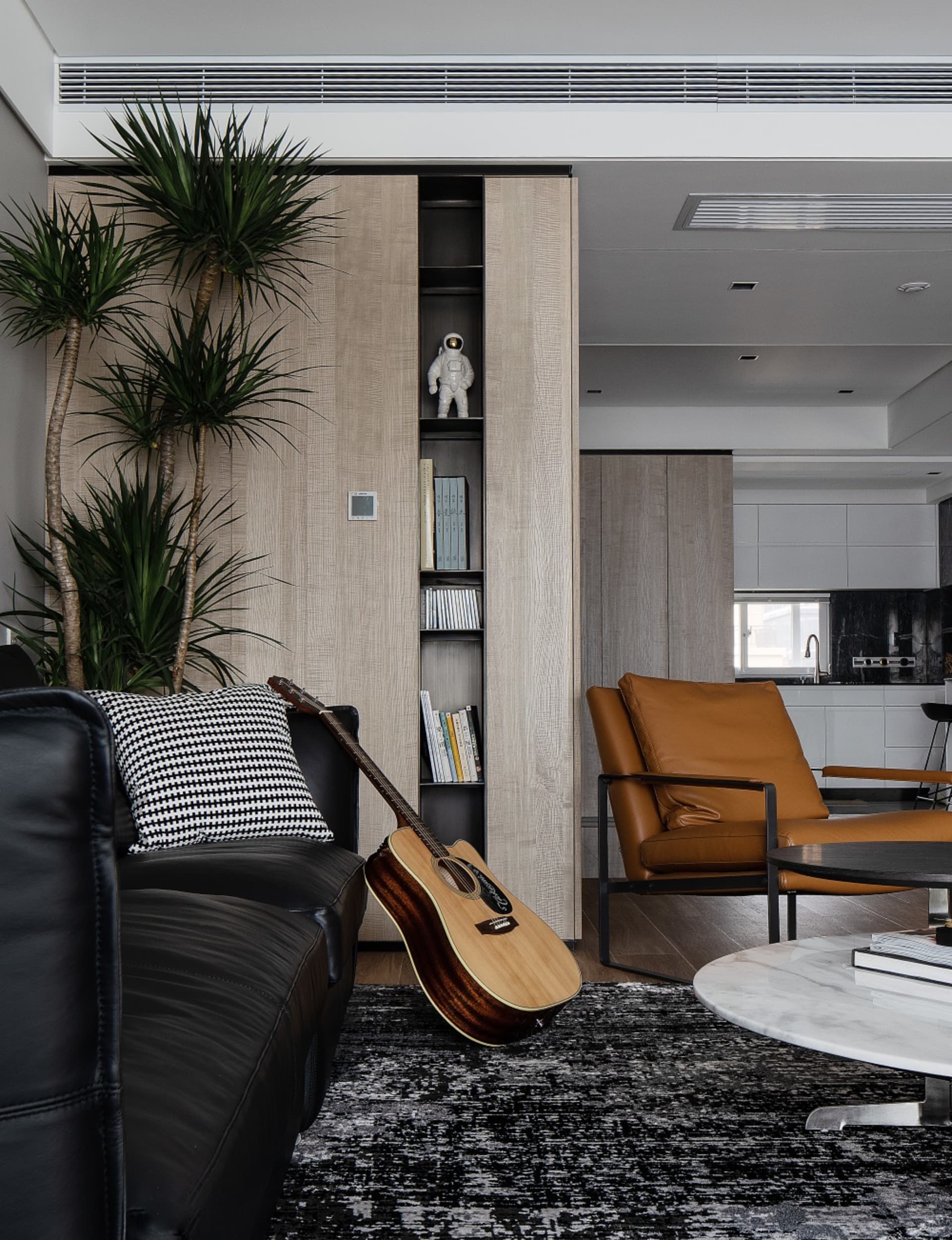 A modern living room with a wooden accent wall, a black leather couch, a brown leather armchair, and a guitar leaning against
