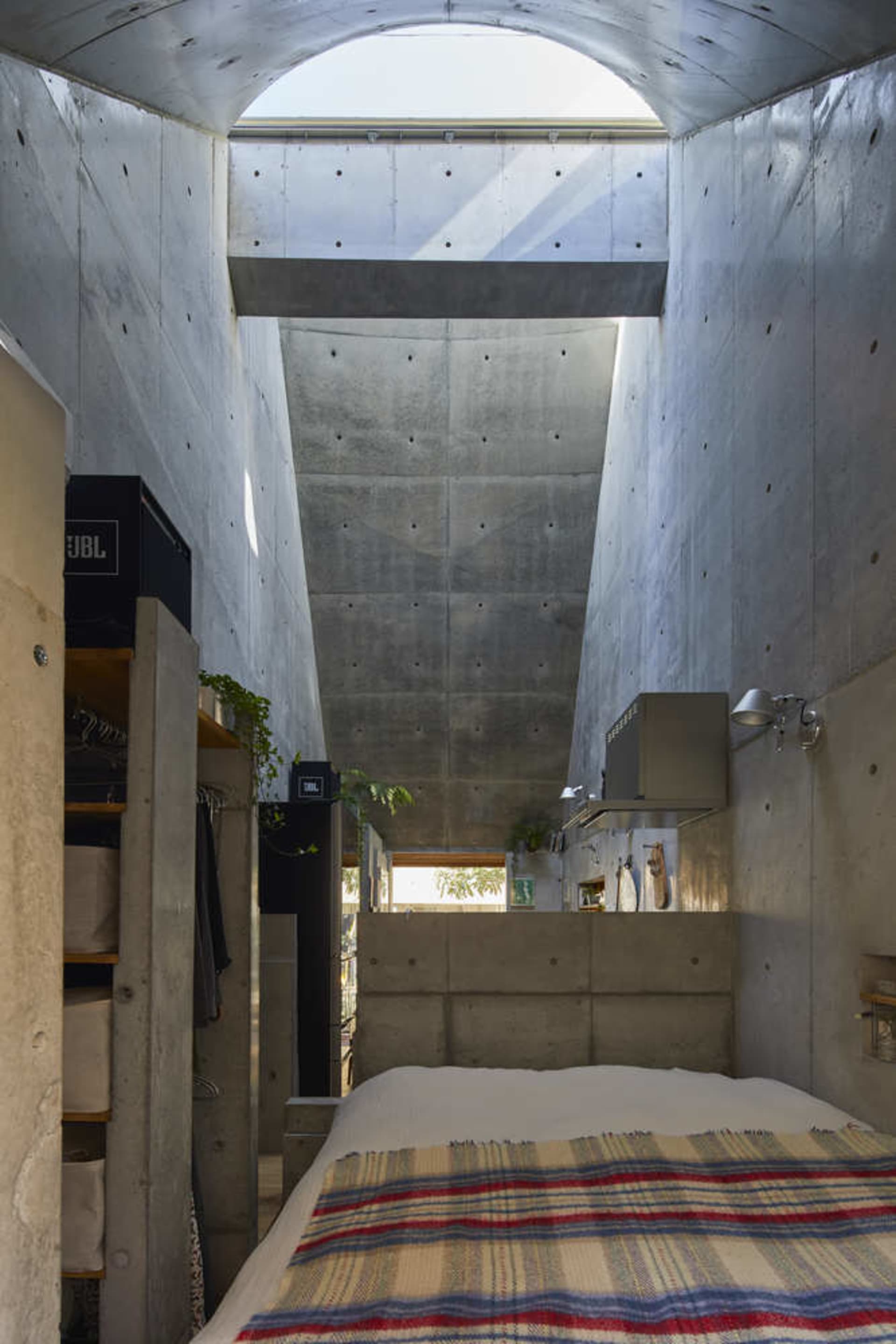 A minimalist bedroom featuring a concrete wall and floor, with a bed covered in a striped blanket and a skylight above.