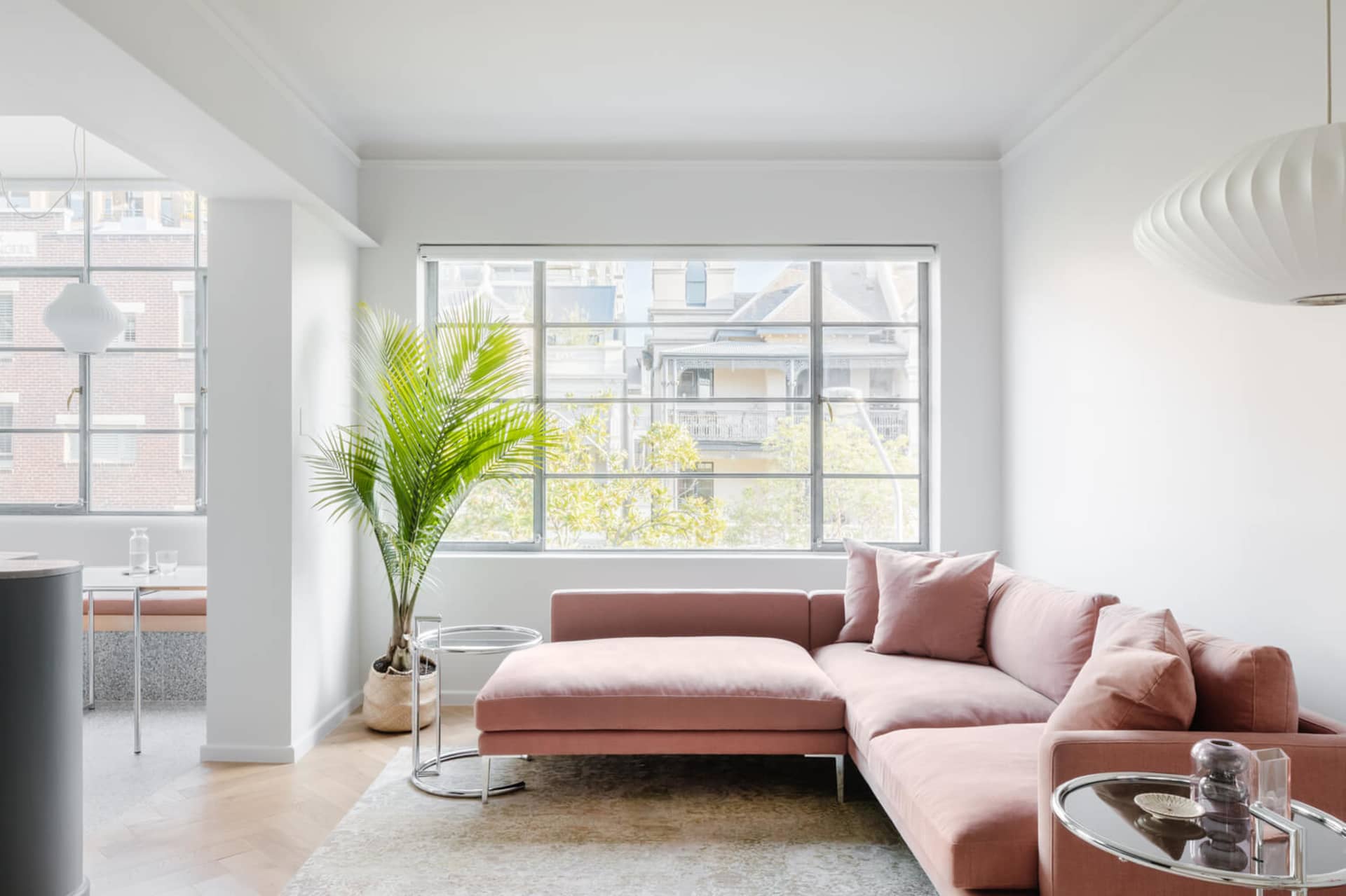 A modern, minimalist living room with a pink sofa, glass table, and large window overlooking urban buildings.