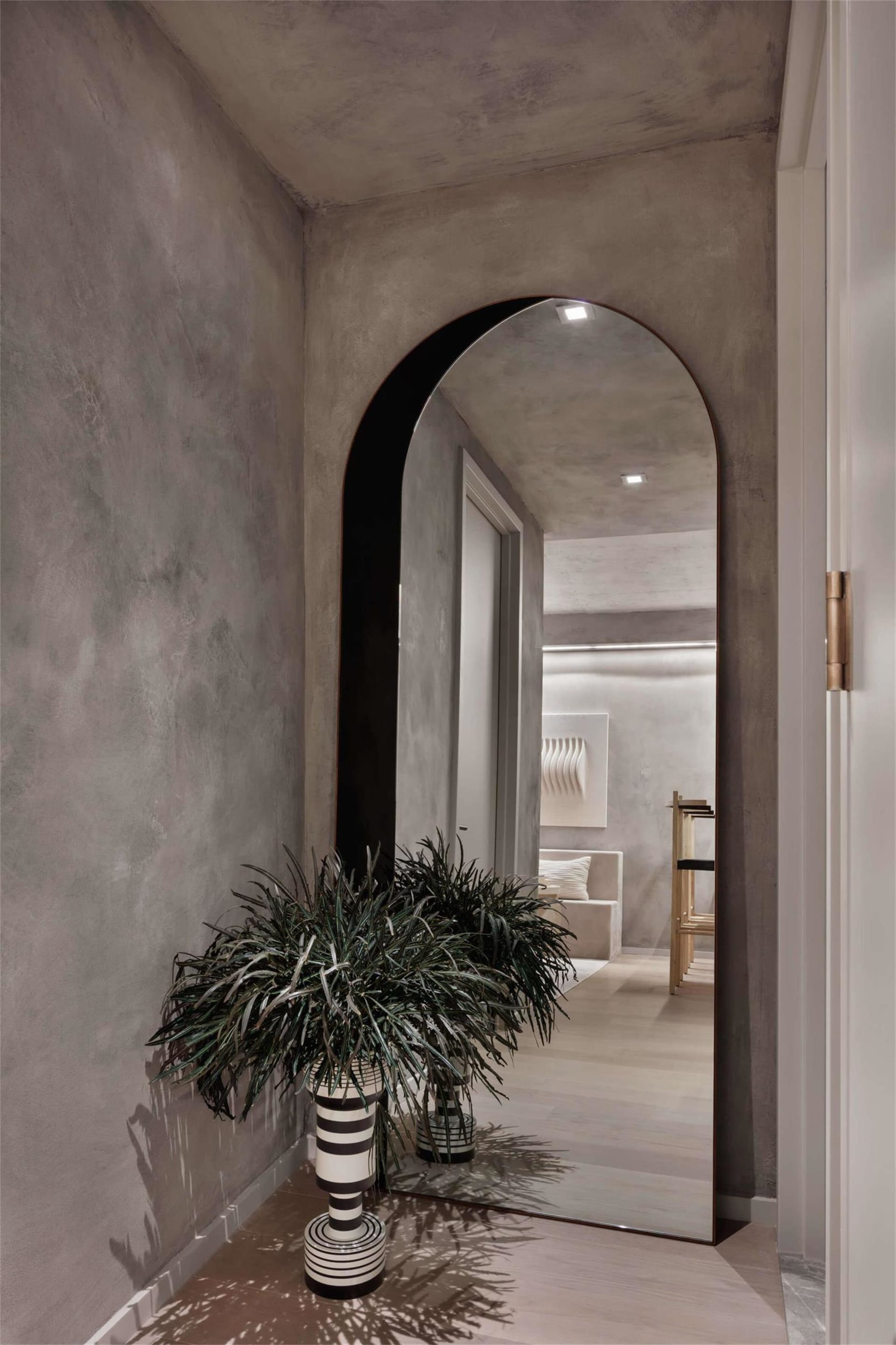A minimalist hallway with a concrete wall and floor, an arched mirror, and a potted plant.