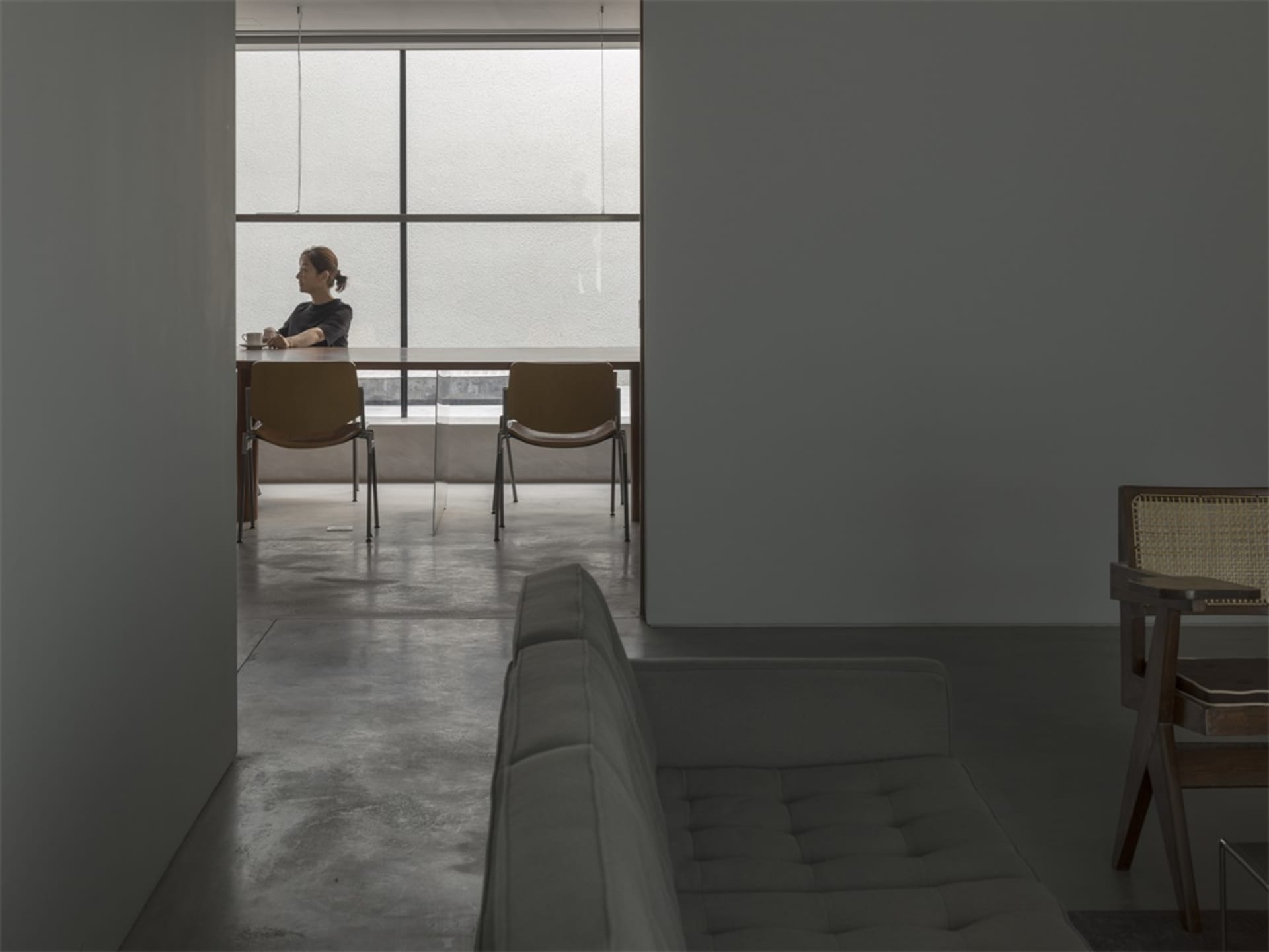 A minimalist modern coffee shop with concrete flooring, a long table, and a woman drinking coffee by a large window.