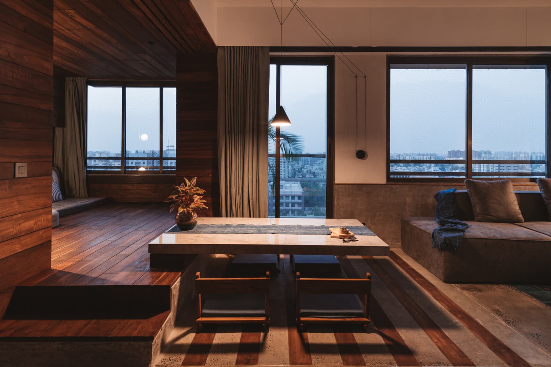 A modern residential lounge featuring wooden seating, concrete flooring, and large glass windows with a cityscape view.