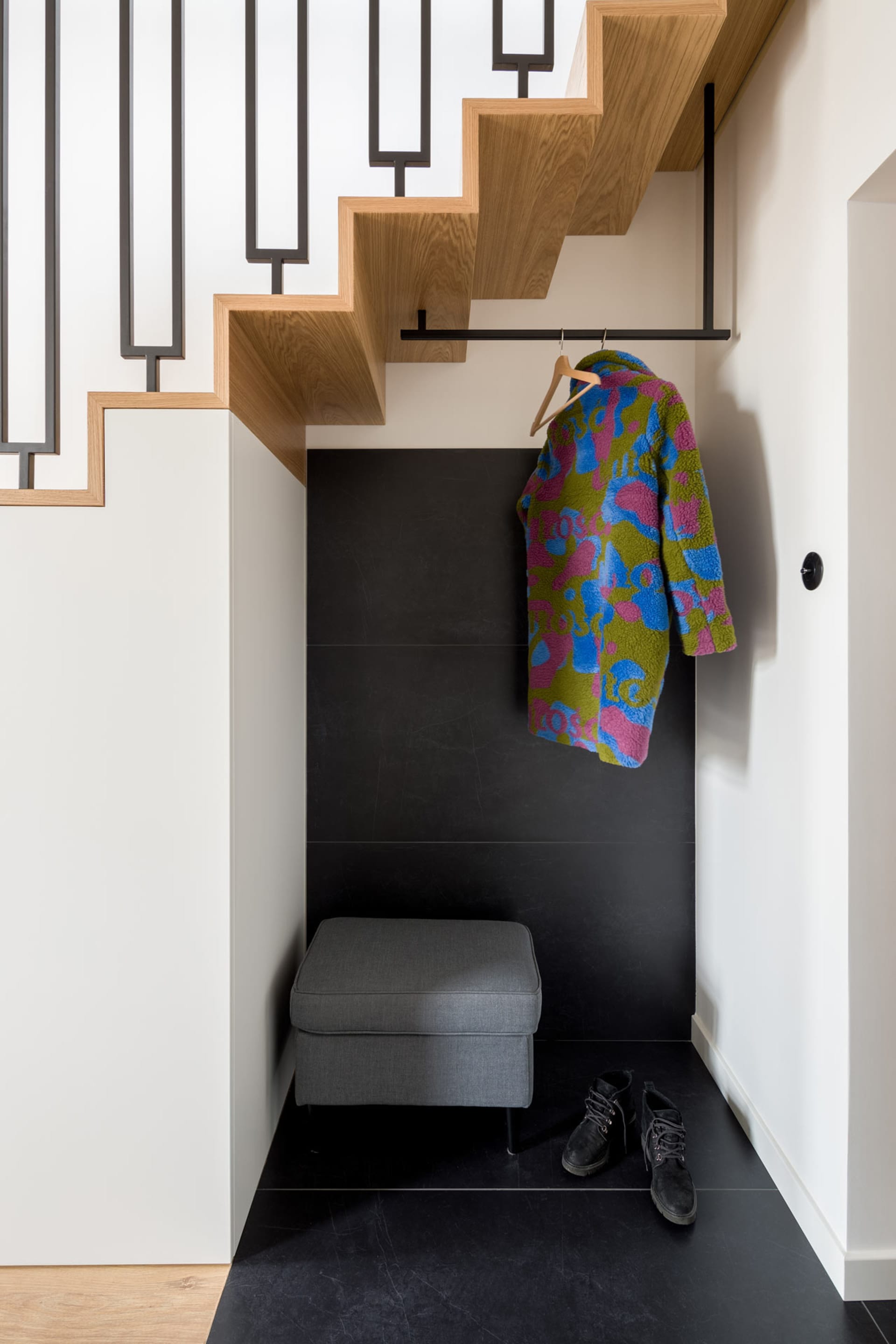 A modern entryway featuring a wooden staircase with a black railing and a colorful coat hanging on a hook.