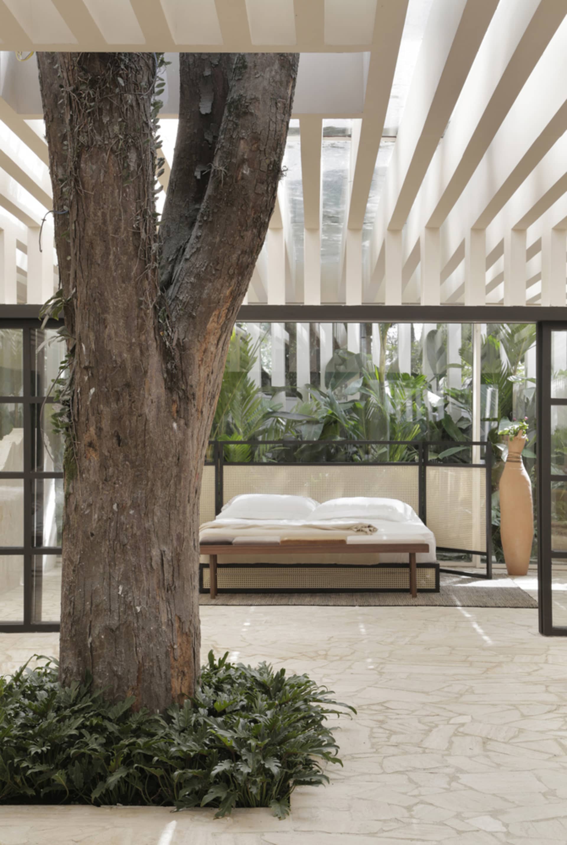 A minimalist bedroom with a white bed and natural wooden elements surrounded by greenery and a tree trunk.