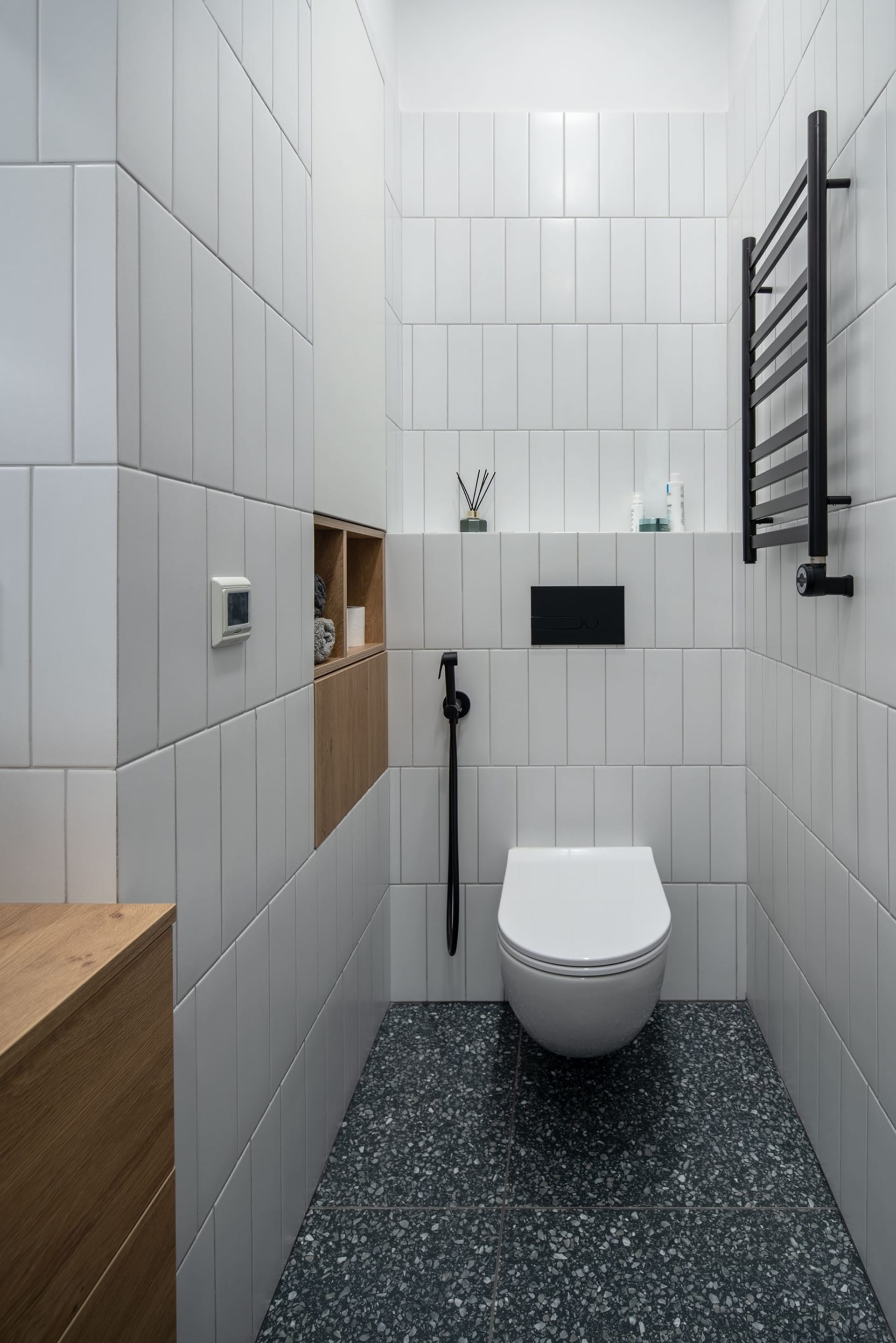 A modern bathroom with white tiles and black terrazzo flooring, featuring a wall-mounted toilet with a black cistern, wooden
