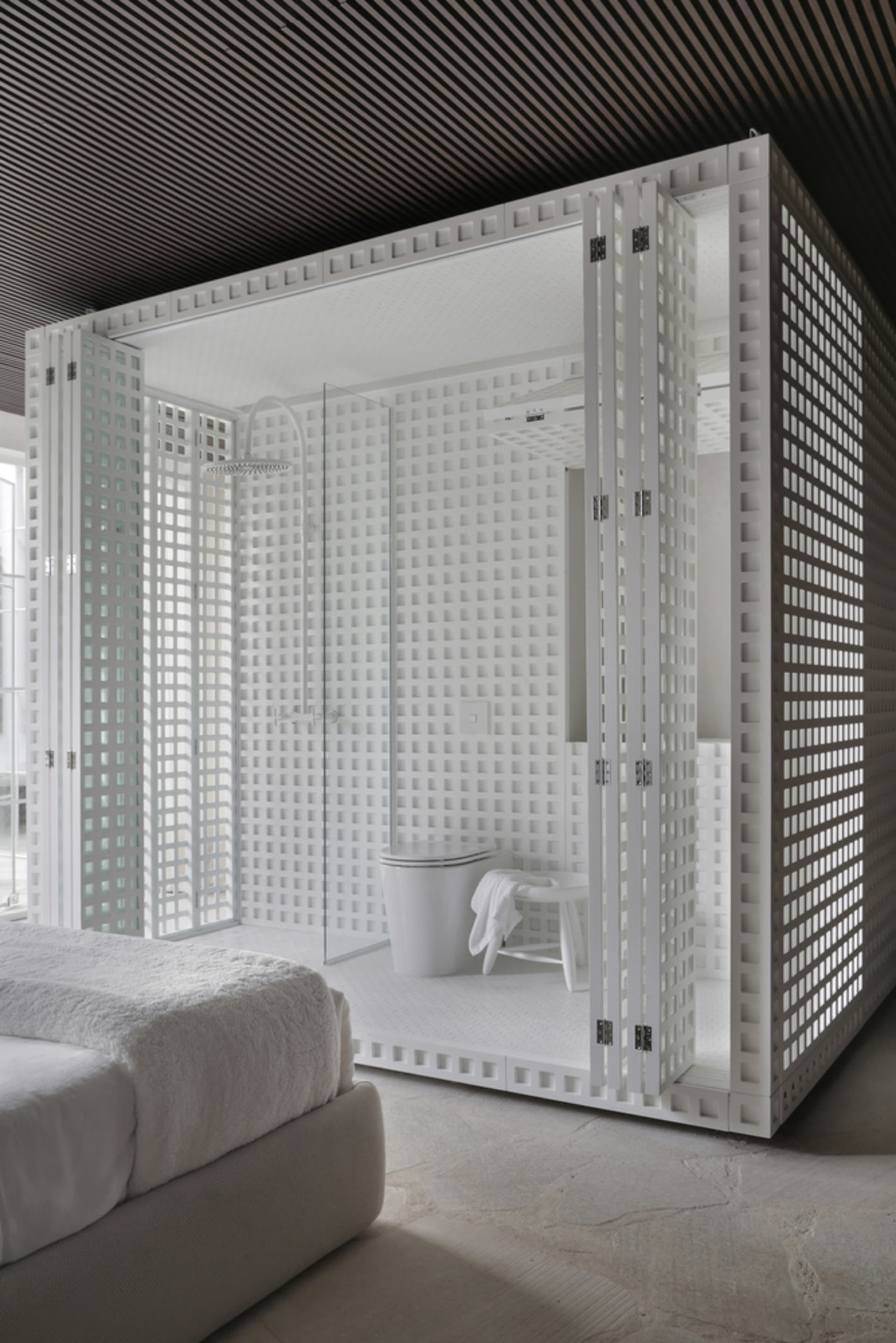 A modern bathroom with a white plaster shower enclosure and a white bed in the foreground.